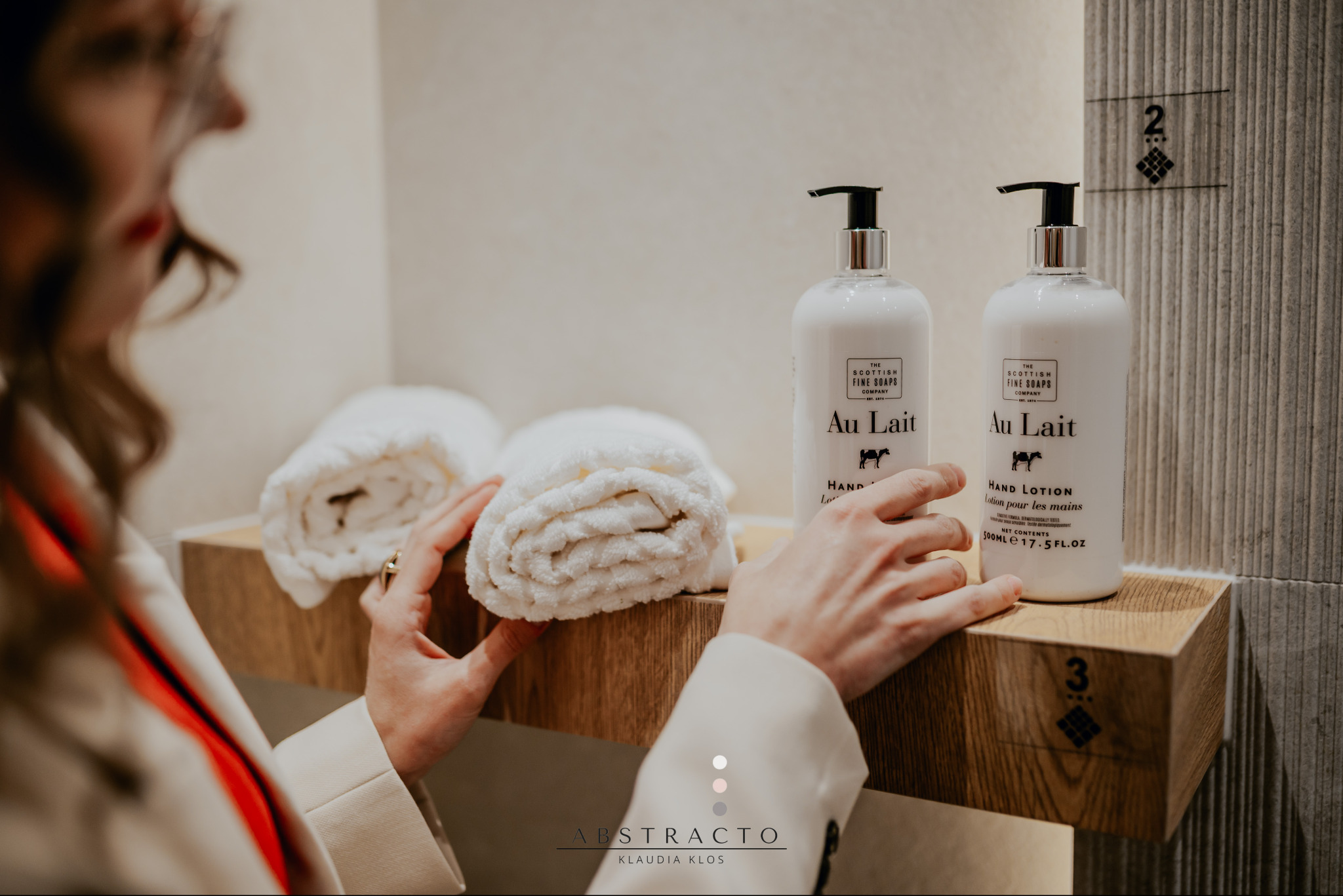 Nowoczesna łazienka z drewnianą półką, na której leżą zwinięte białe ręczniki i butelki z balsamem do rąk Au Lait. Kobieta układa ręczniki na półce.