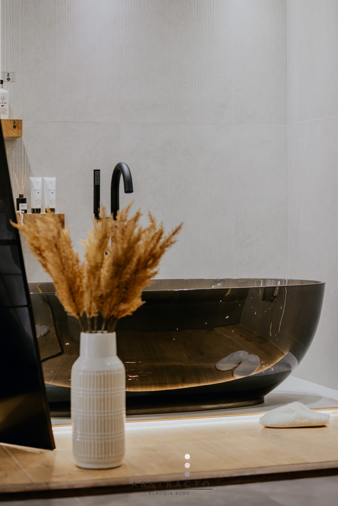 Nowoczesna łazienka z owalną, ciemną wanną wolnostojącą, czarną armaturą i dekoracyjnym wazonem z suszonymi trawami. Minimalistyczny design, ciepłe oświetlenie LED.