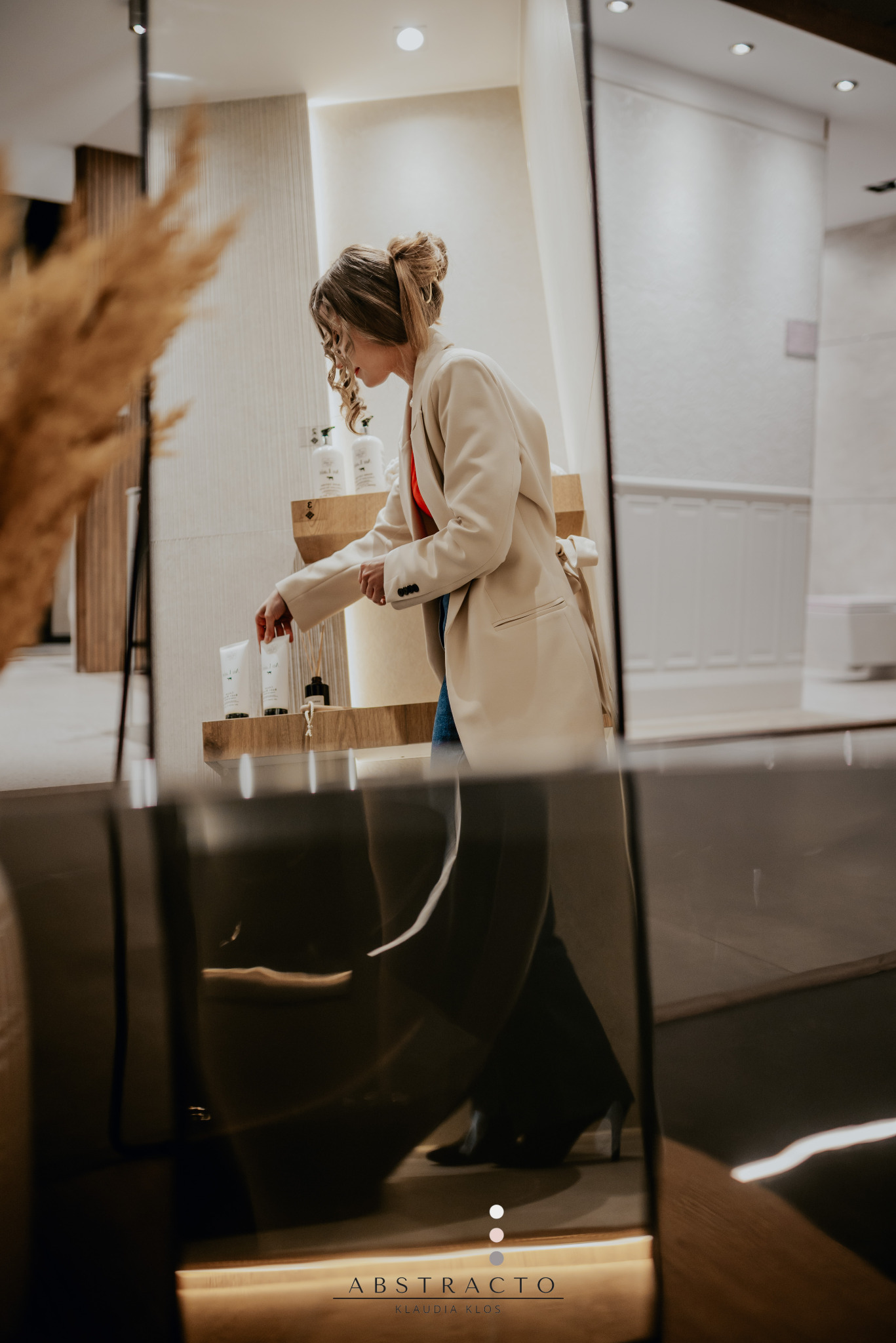 Kobieta w beżowym żakiecie przegląda kosmetyki na drewnianej półce w jasnym wnętrzu. Ujęcie zza szklanej tafli.