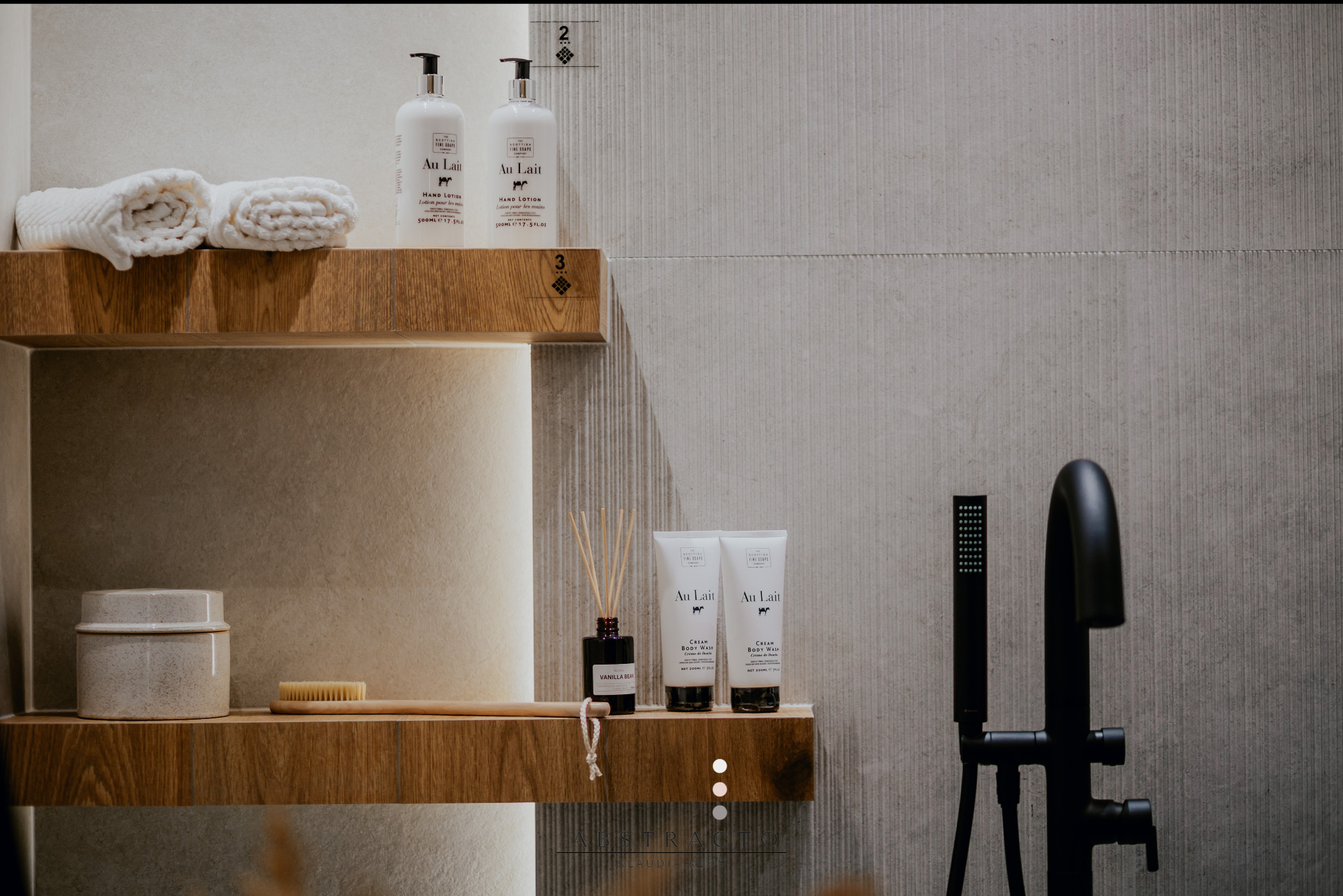 Minimalistyczna łazienka z drewnianymi półkami, na których stoją kosmetyki Au Lait i ręczniki. Czarna bateria umywalkowa na tle szarej, teksturowanej ściany.