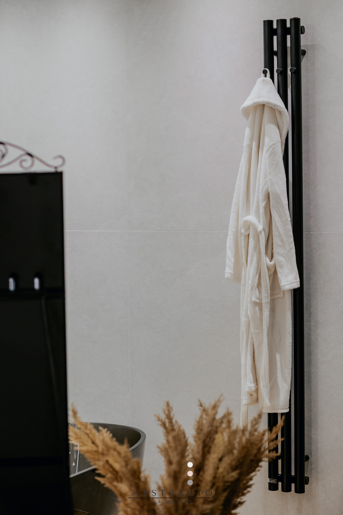 Minimalistyczna łazienka z czarnym grzejnikiem drabinkowym, na którym wisi biały szlafrok. U dołu dekoracyjne trawy i fragment umywalki.