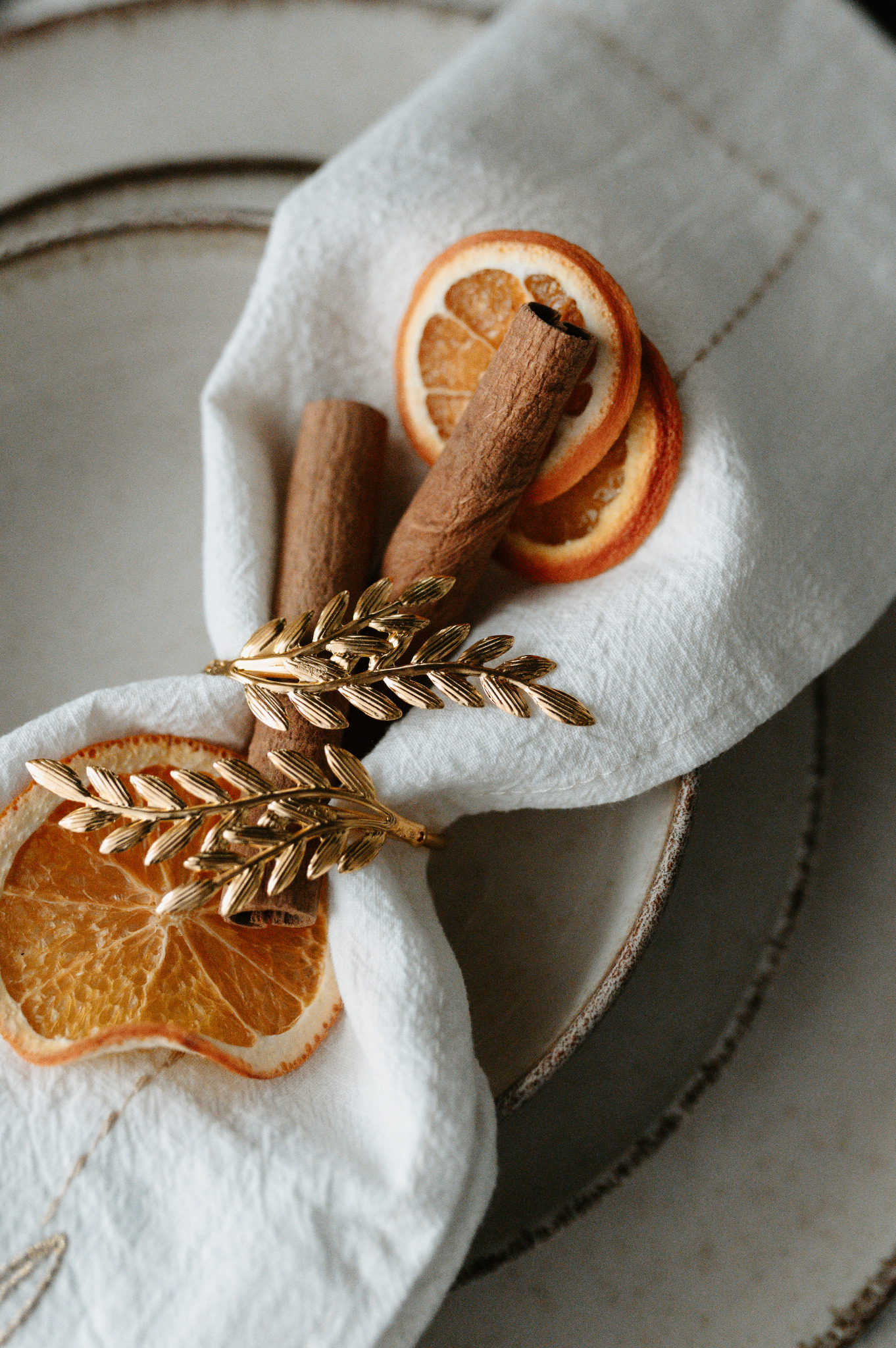 Dekoracja stołu: biała serwetka z pierścieniem w kształcie gałązki, suszone pomarańcze i laski cynamonu na talerzu. Ciepłe, naturalne światło.
