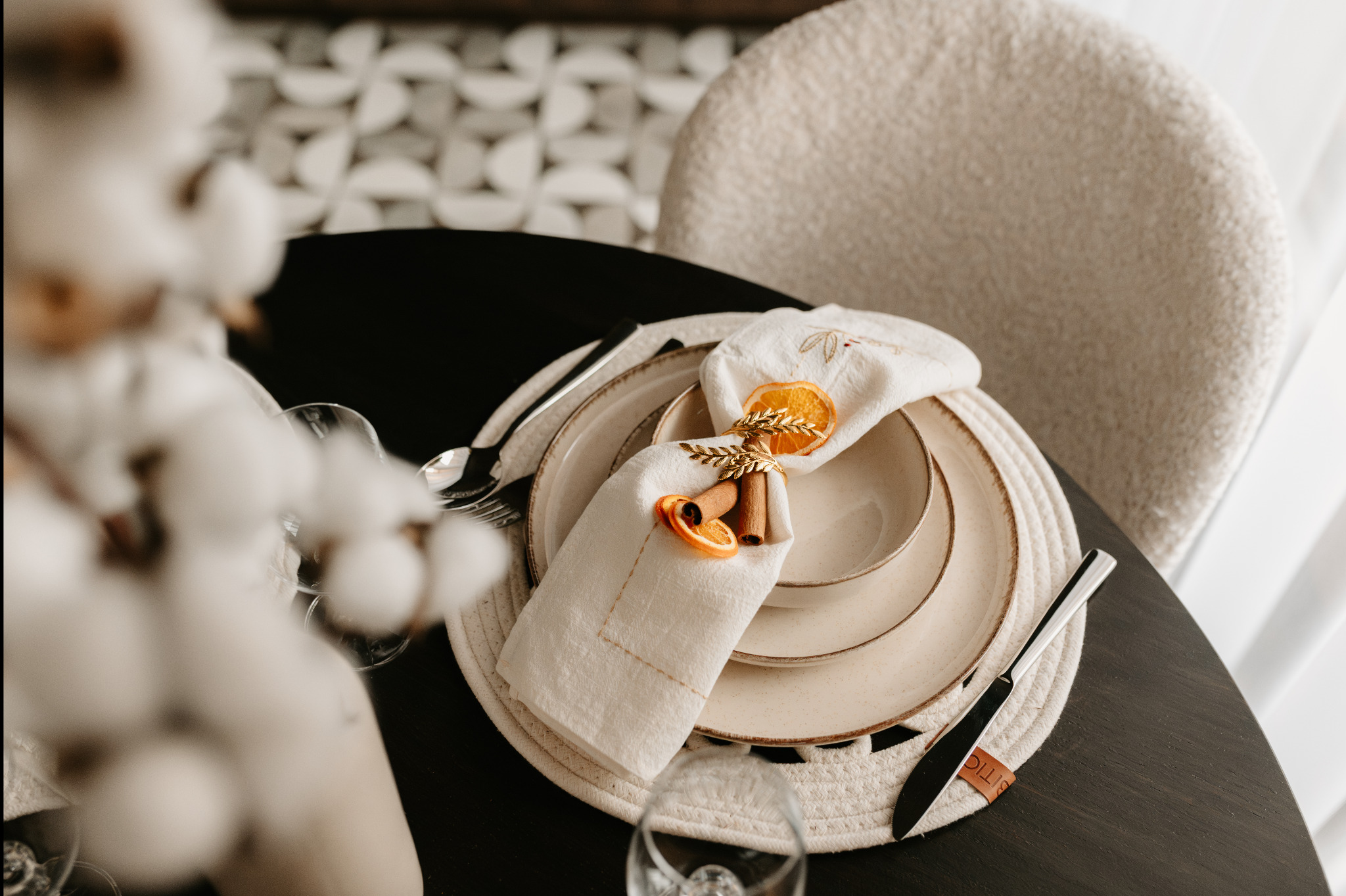 Elegancko nakryty stół z beżową zastawą, lnianą serwetką zdobioną suszonymi pomarańczami i cynamonem, na tle geometrycznej ściany i krzesła w stylu bouclé.