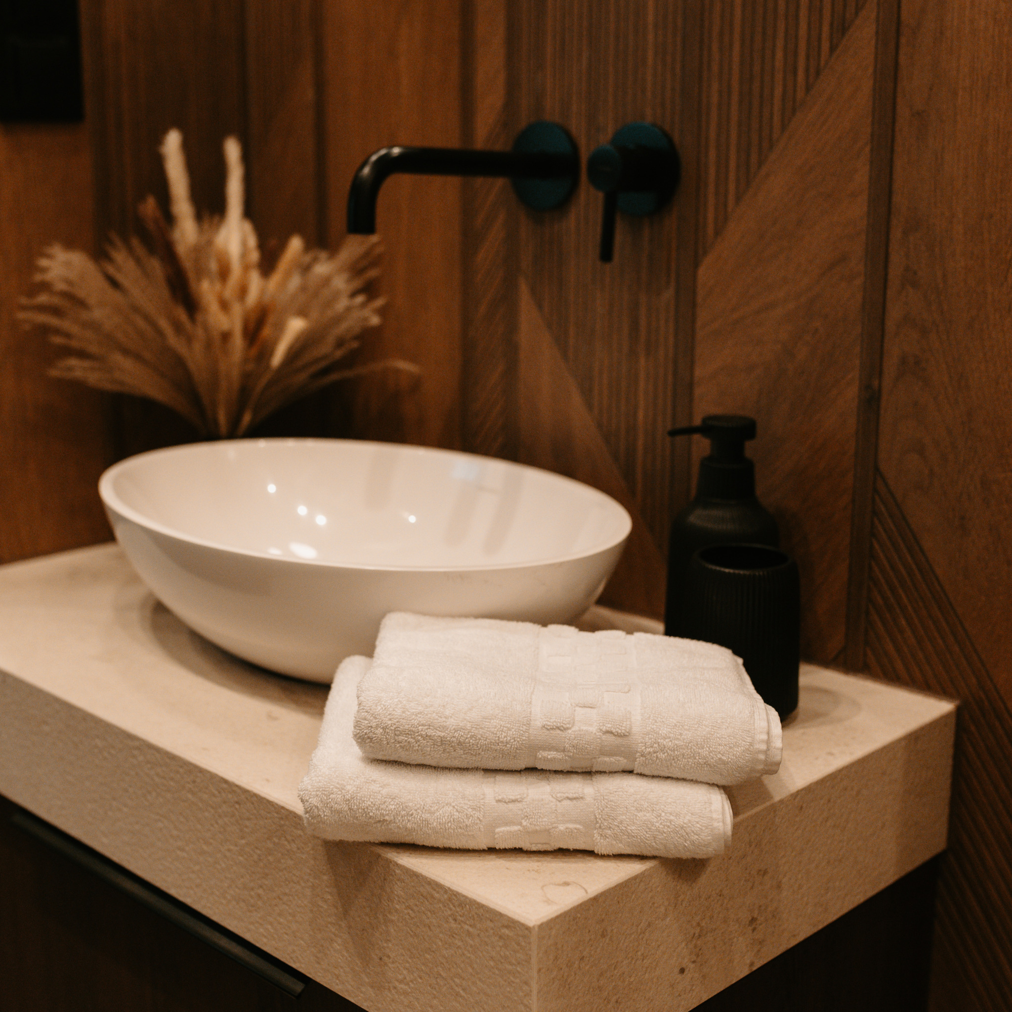 Nowoczesna łazienka: biała umywalka nablatowa, czarna bateria i dozownik, beżowy blat z ręcznikami na tle drewnianej ściany. Minimalistyczny design.