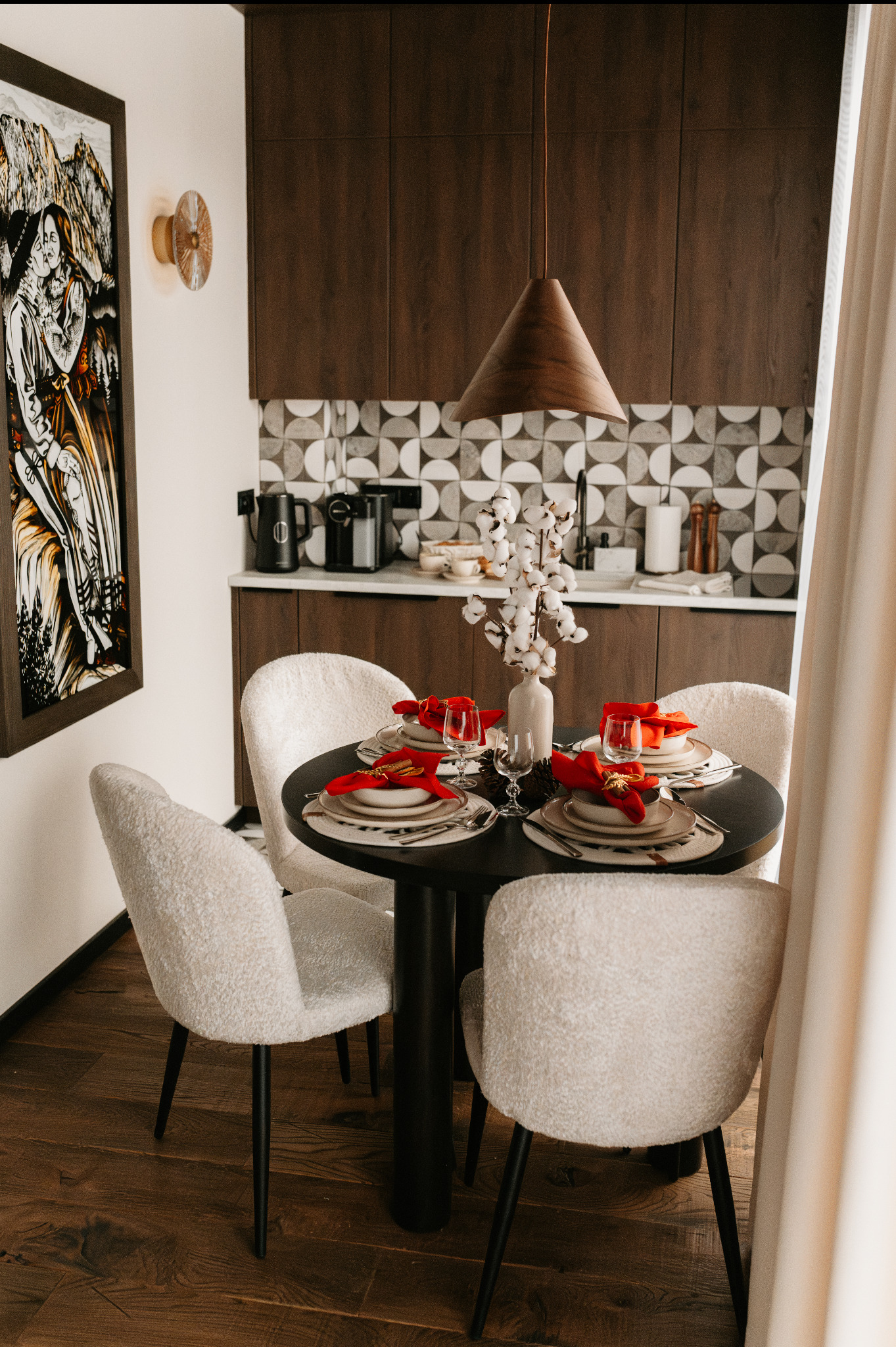 Stylowa jadalnia z okrągłym, czarnym stołem i czterema krzesłami z jasną tapicerką. Na stole dekoracje i zastawa w odcieniach bieli i czerwieni. W tle kuchnia z ciemnymi szafkami.