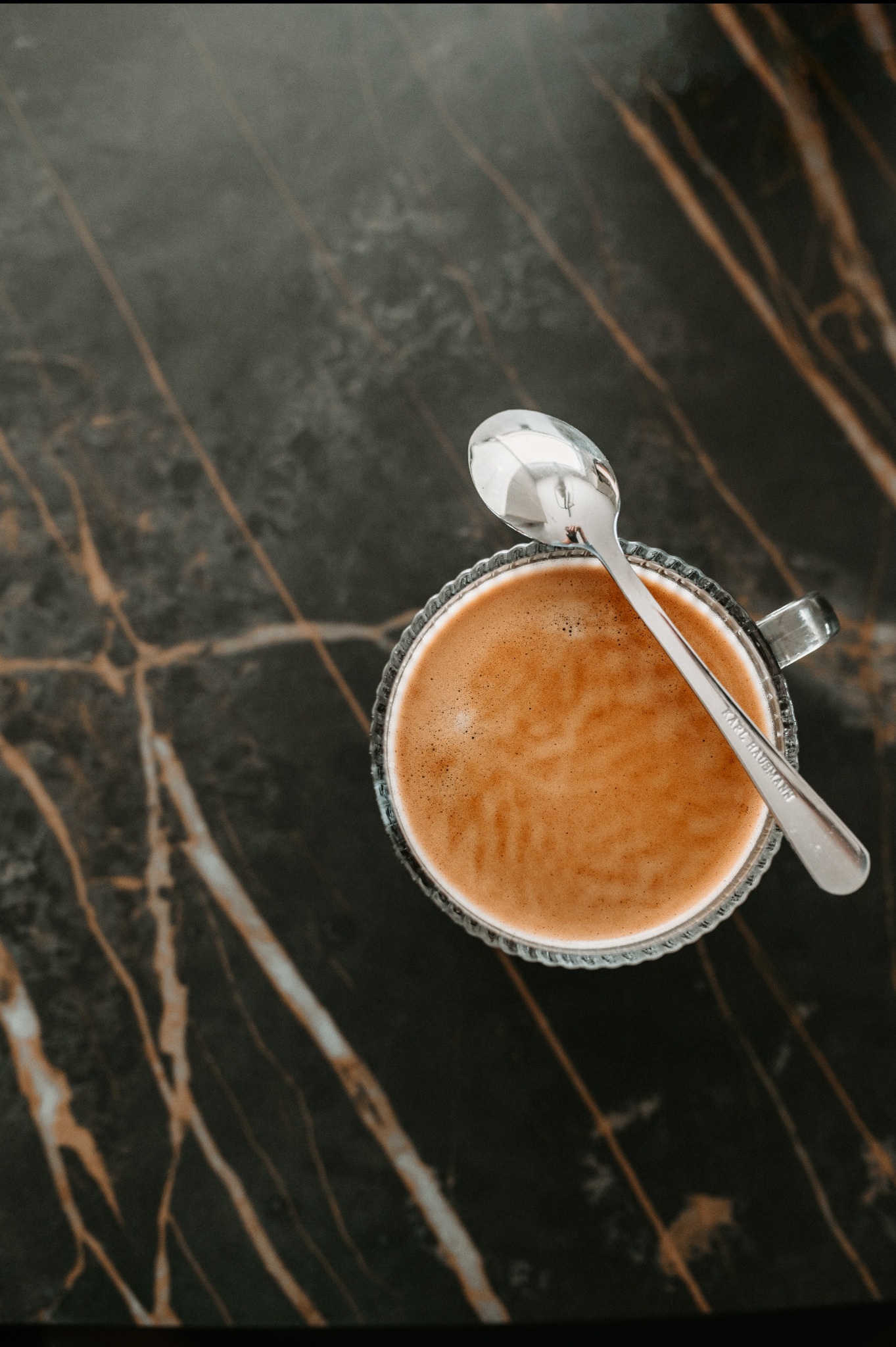 Kawa w szklance z łyżeczką na marmurowym blacie. Minimalistyczne ujęcie z góry, podkreślające teksturę kamienia i ciepły odcień napoju.