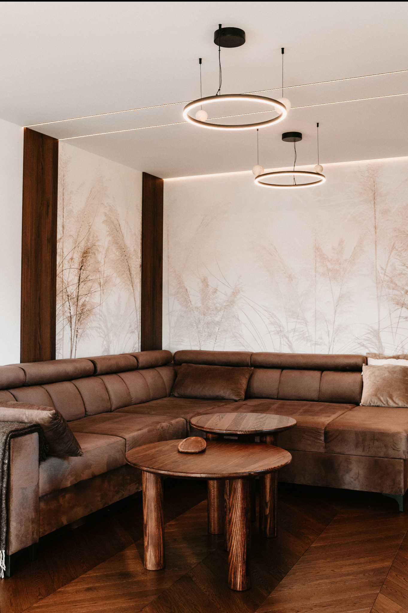 Stylowy salon z brązową, narożną sofą, drewnianymi stolikami kawowymi i dekoracyjną tapetą z motywem traw. Oświetlenie stanowią okrągłe lampy wiszące.