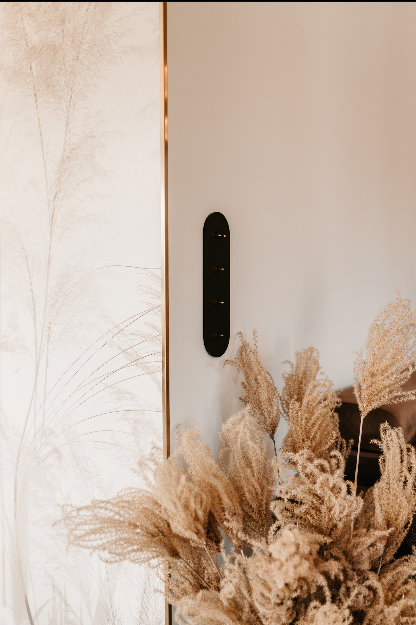 Beżowe wnętrze z tapetą w trawy, czarnym wieszakiem na ścianie i bukietem suszonych traw. Minimalistyczny design z akcentami natury.