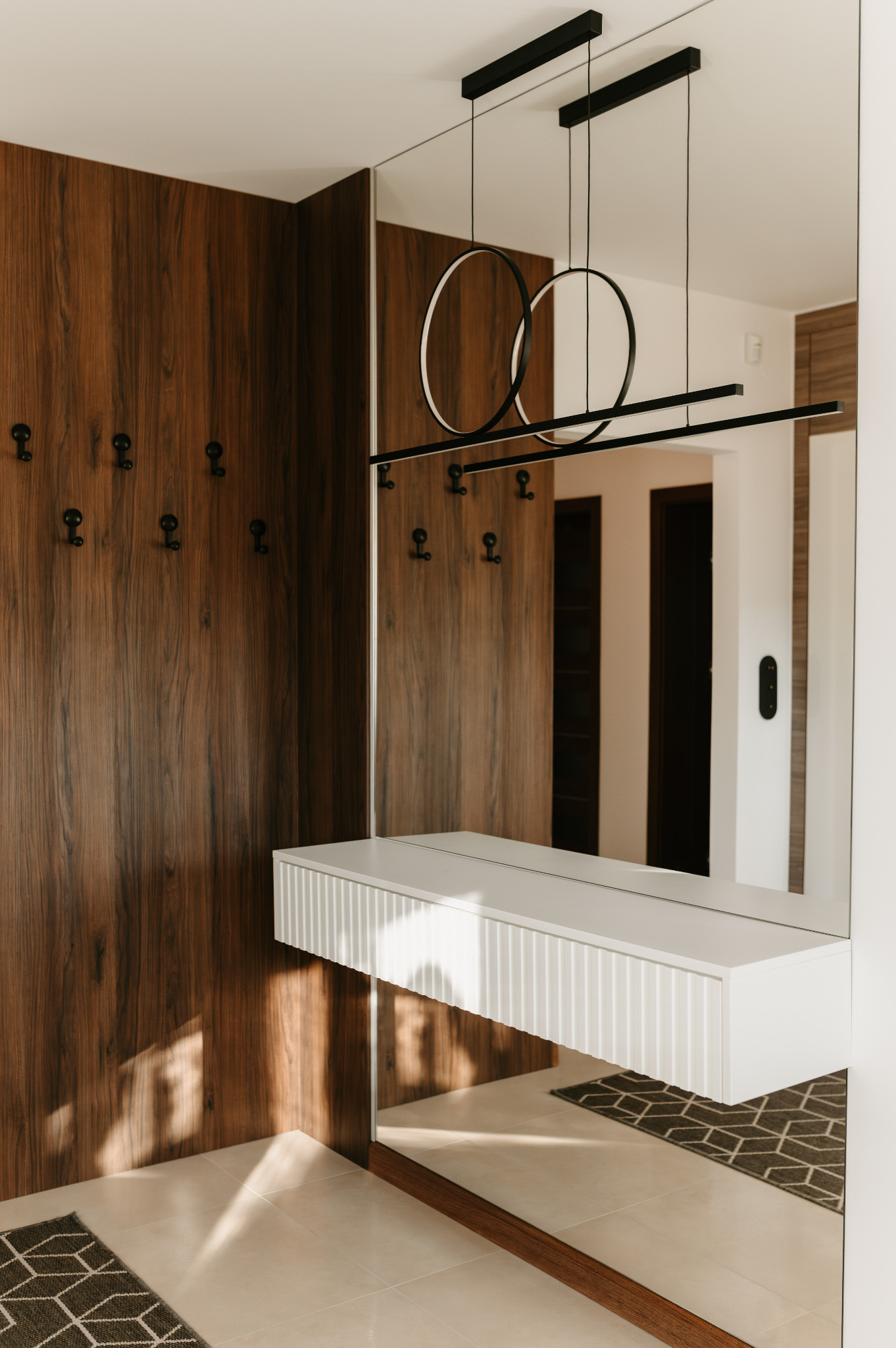 Nowoczesny przedpokój z drewnianą zabudową, lustrem i minimalistycznym oświetleniem. Widoczny fragment dywanu z geometrycznym wzorem i biała konsola.