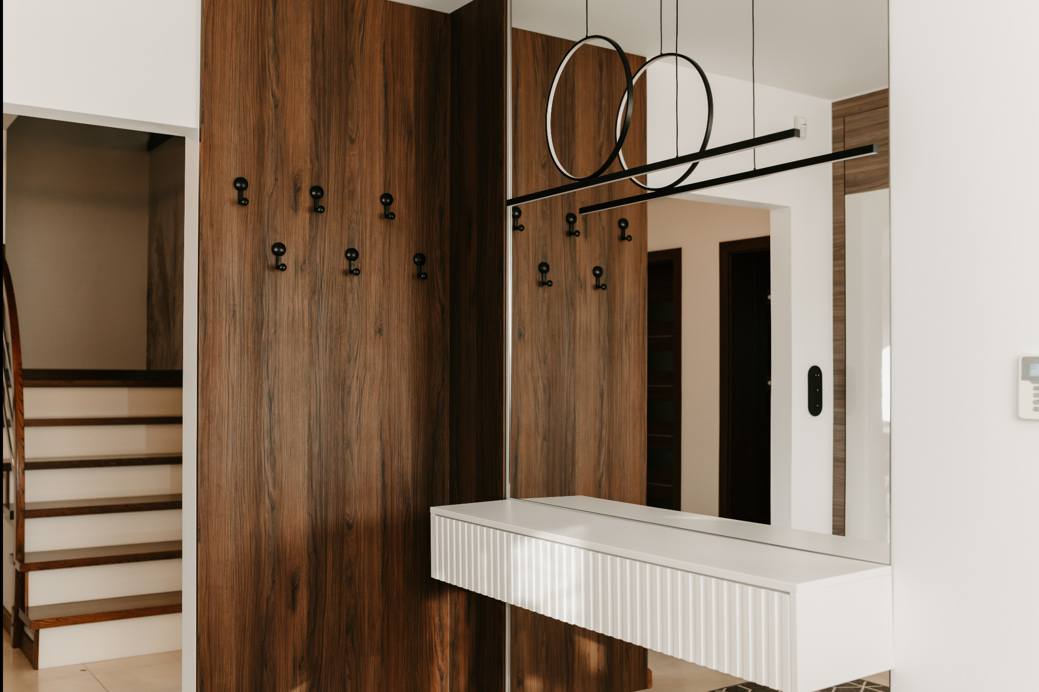 Nowoczesny przedpokój z drewnianą zabudową, wieszakami i lustrem. Minimalistyczna lampa sufitowa. Widoczne schody i wejście do innego pomieszczenia.