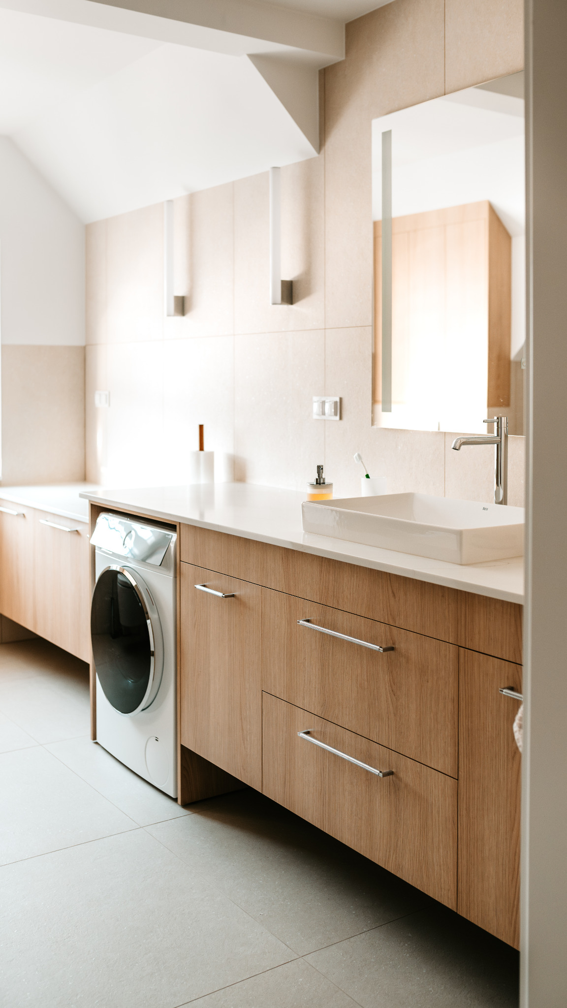 Jasna łazienka z drewnianymi szafkami, białą umywalką i pralką w zabudowie. Minimalistyczny design, beżowe płytki, lustro z oświetleniem.