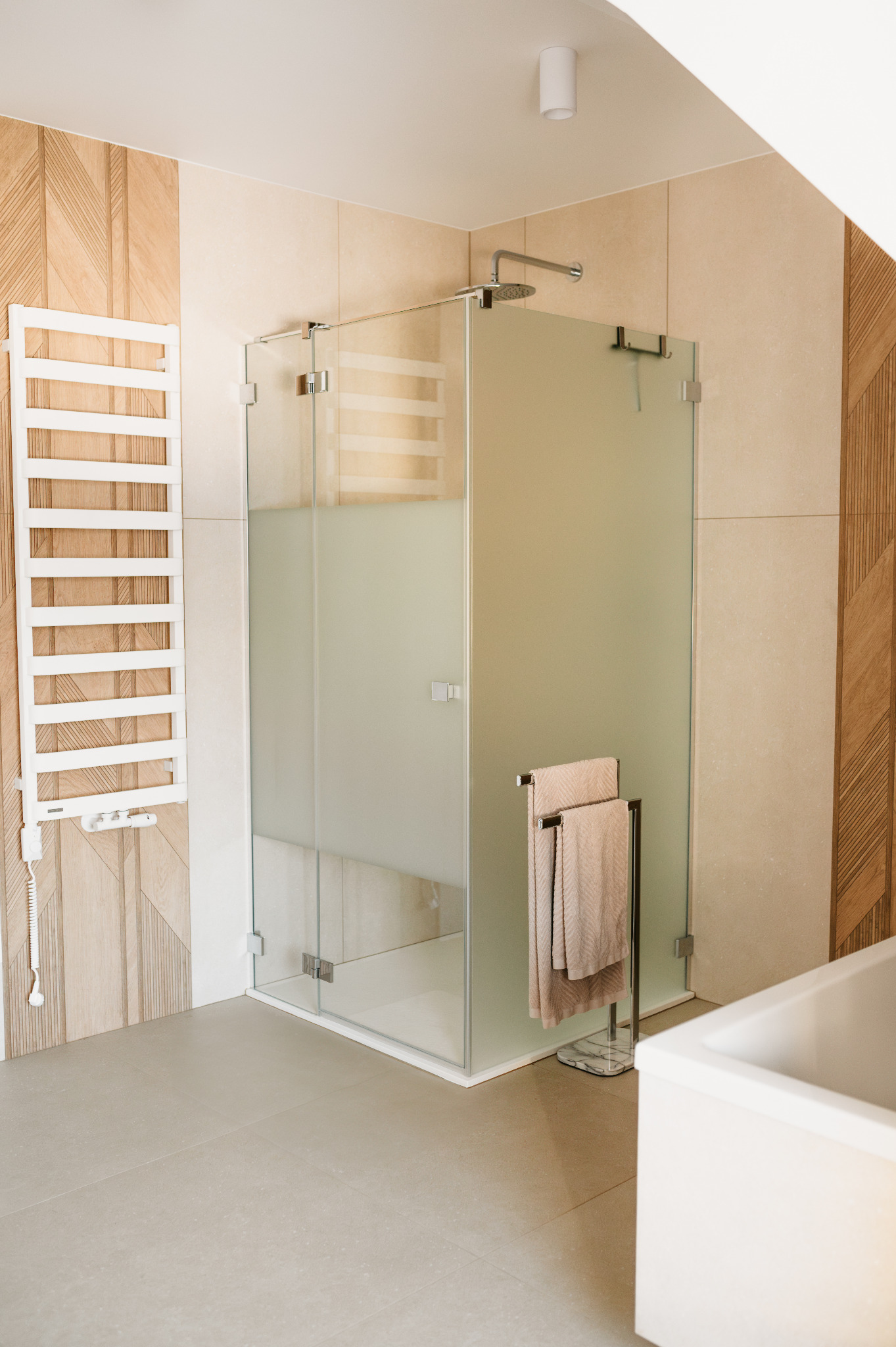 Nowoczesna kabina prysznicowa z matowego szkła, ręczniki na wieszaku, grzejnik drabinkowy i drewniane panele ścienne w beżowej łazience. Minimalistyczny design.