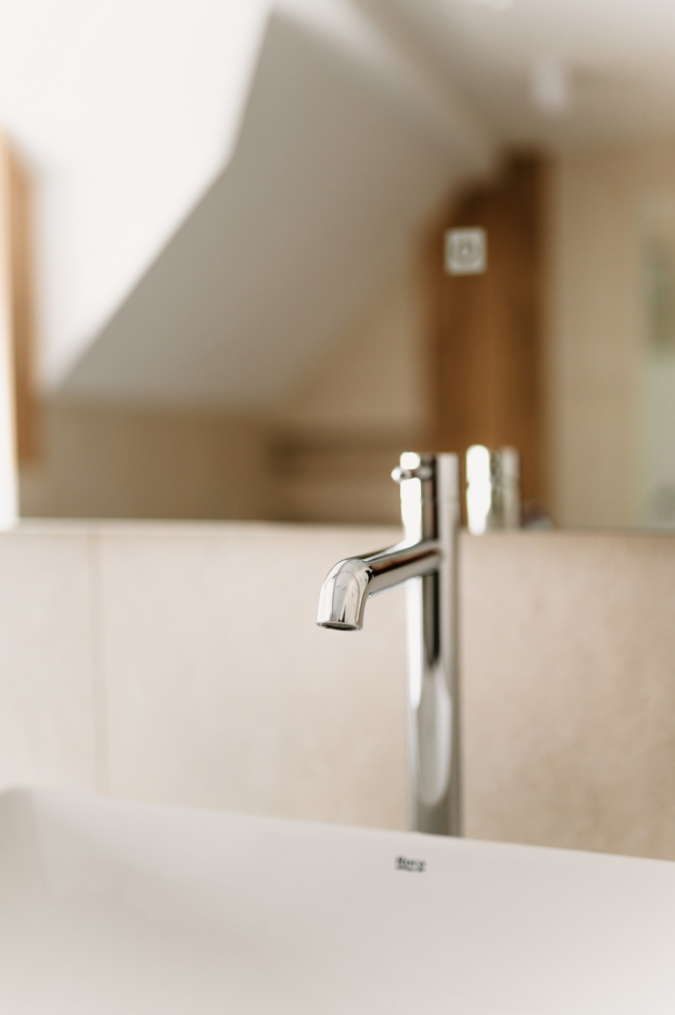 Błyszcząca, chromowana bateria umywalkowa na tle beżowych płytek i lustra w łazience. Minimalistyczny design, ciepłe oświetlenie, nowoczesne wykończenie.