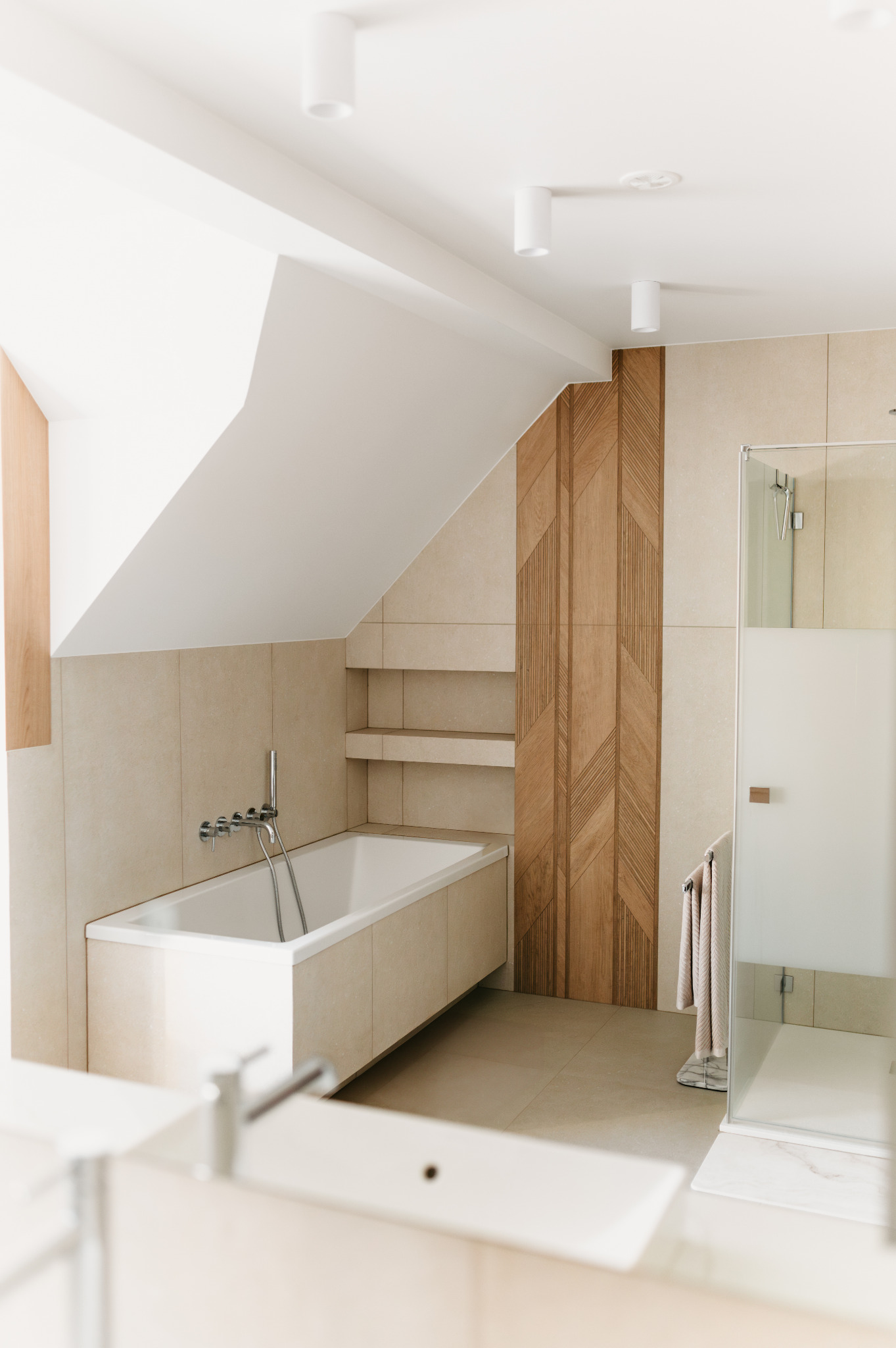 Nowoczesna łazienka na poddaszu w Rabce-Zdroju. Beżowe płytki, drewniane panele dekoracyjne, wanna i kabina prysznicowa. Minimalistyczny design i jasne oświetlenie.