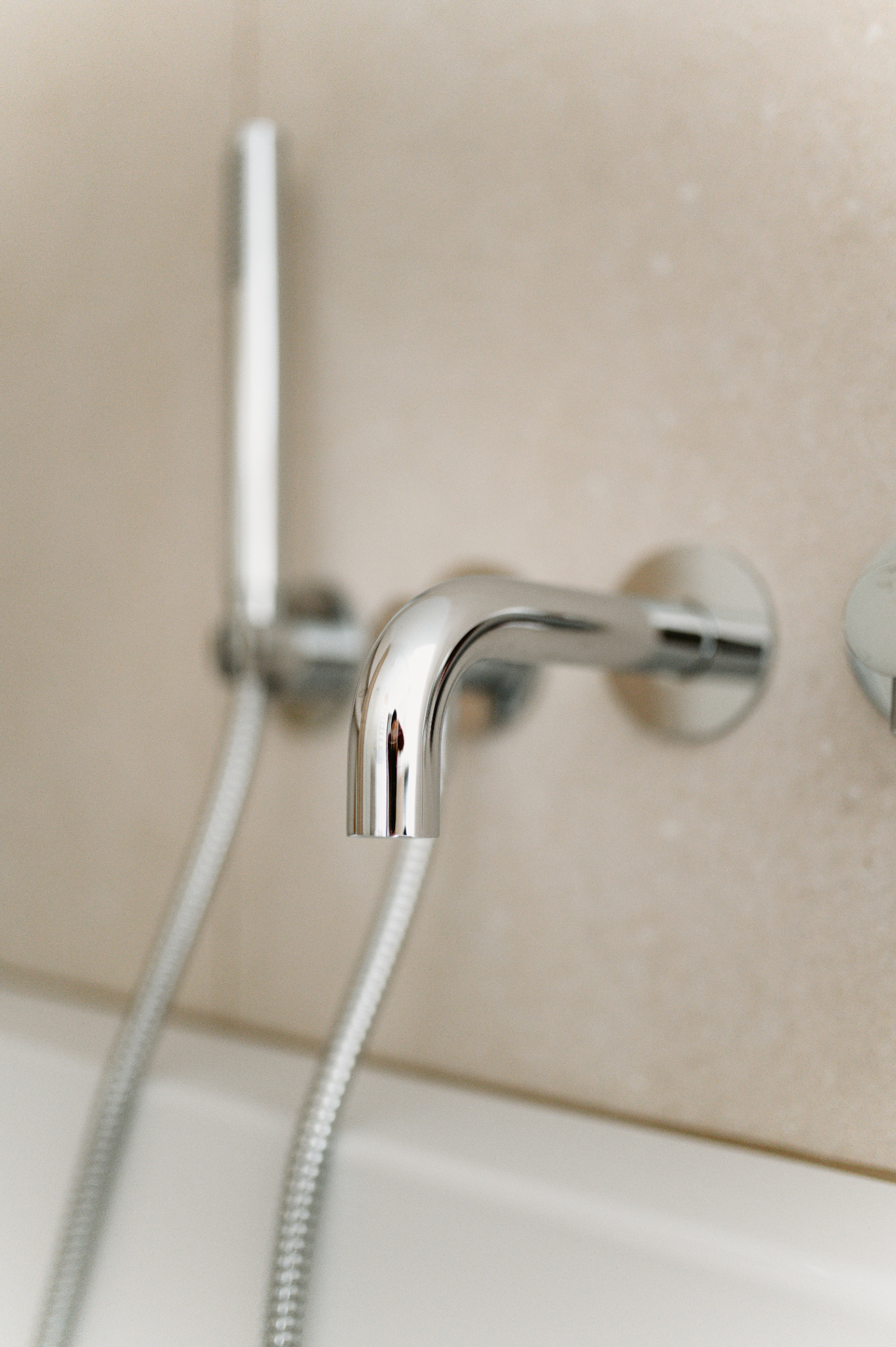 Błyszcząca, chromowana bateria wannowa ścienna z wężem i słuchawką prysznicową na tle beżowej ściany. Minimalistyczny design, nowoczesna armatura łazienkowa.