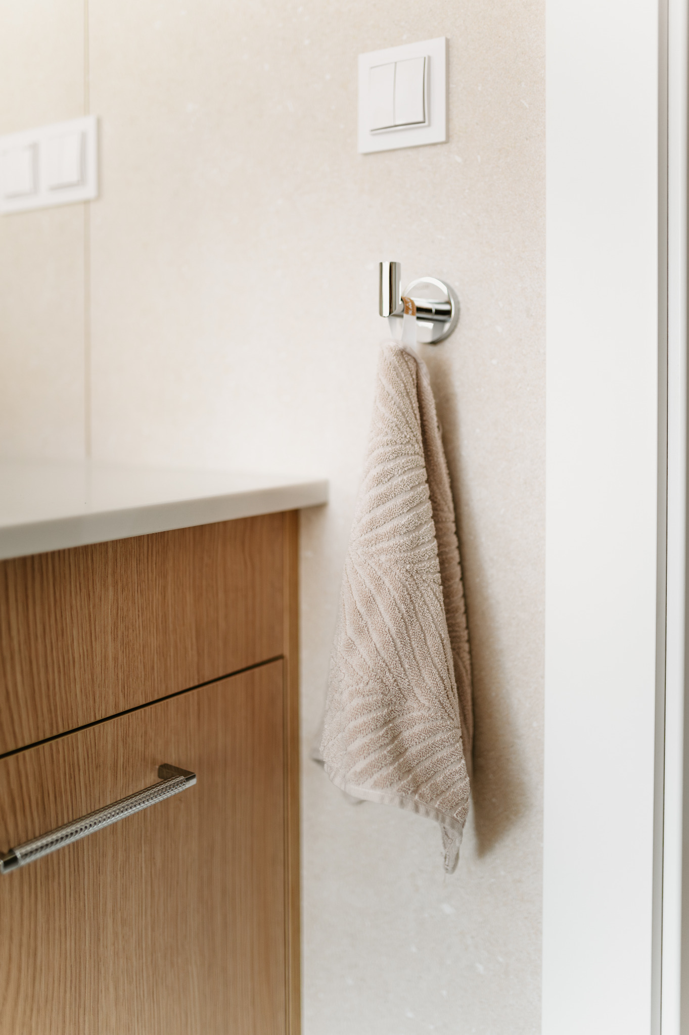 Jasna łazienka: beżowy ręcznik na chromowanym wieszaku, drewniana szafka z blatem i przełącznik światła. Minimalistyczny design, neutralna kolorystyka.