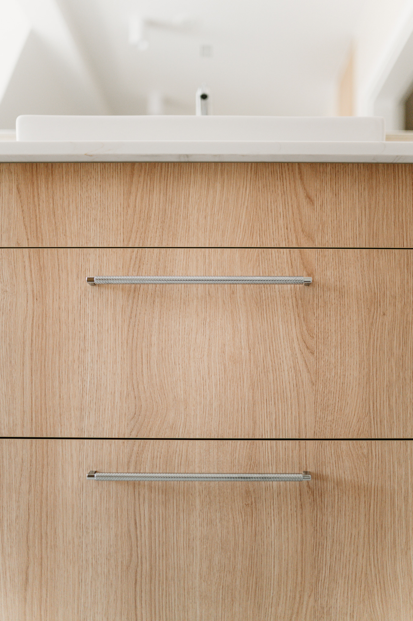 Drewniana szafka łazienkowa z dwoma szufladami i srebrnymi uchwytami, umywalka nablatowa w tle. Minimalistyczny design, jasne oświetlenie.