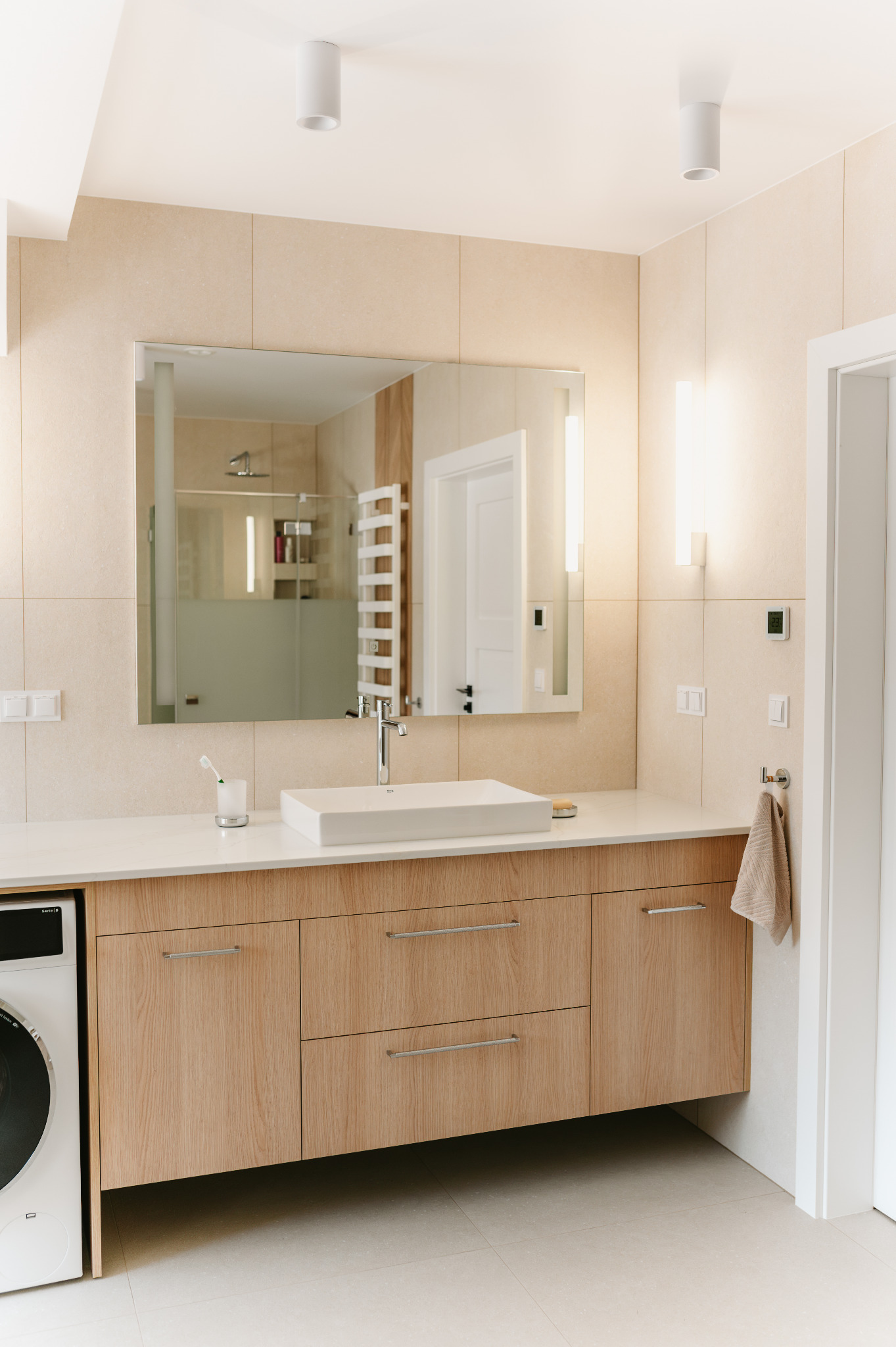 Jasna łazienka z beżowymi płytkami, drewnianą szafką pod umywalką i dużym lustrem. Widoczna pralka w zabudowie i minimalistyczne oświetlenie.