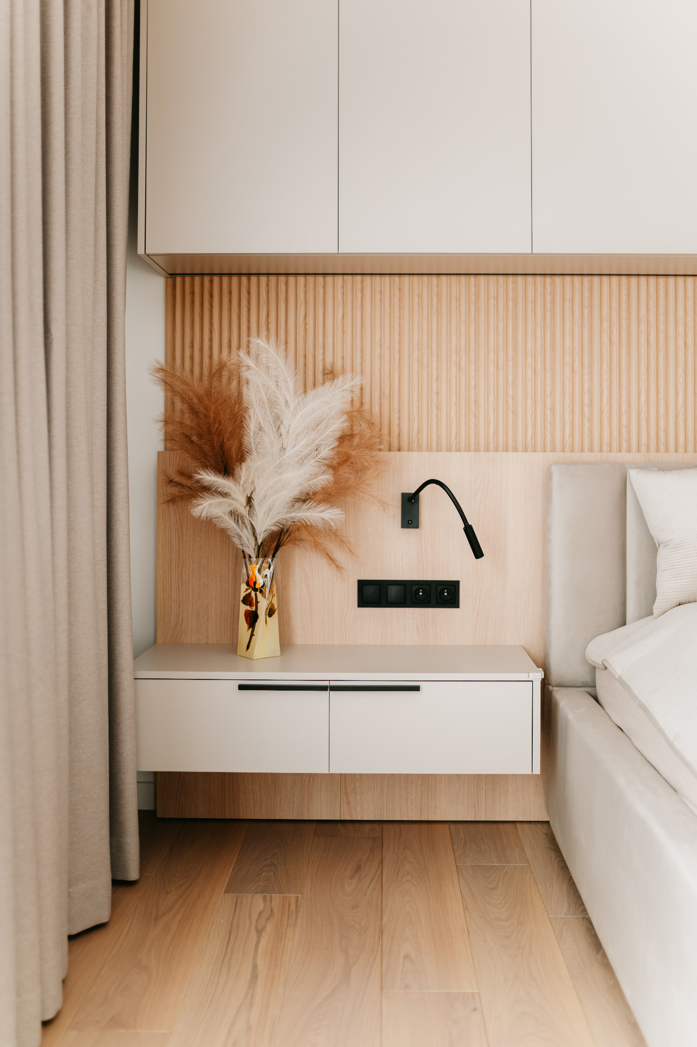 Nowoczesna sypialnia: drewniana ściana z panelem, biała szafka nocna z czarnymi uchwytami, lampka i wazon z trawami. Parkiet na podłodze, beżowa zasłona z lewej.