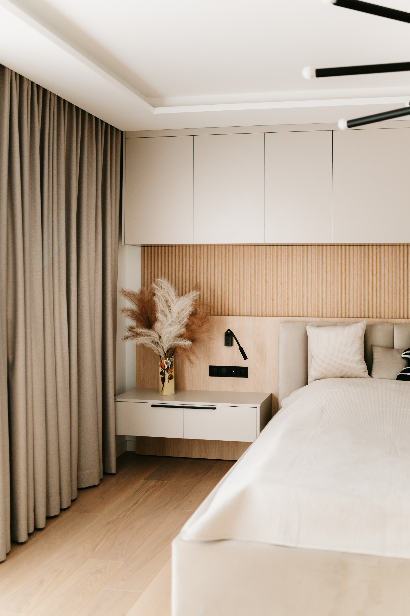 Jasna sypialnia z beżowymi zasłonami i drewnianą ścianą za łóżkiem. Na szafce nocnej wazon z pampasową trawą i lampka. Minimalistyczny design.