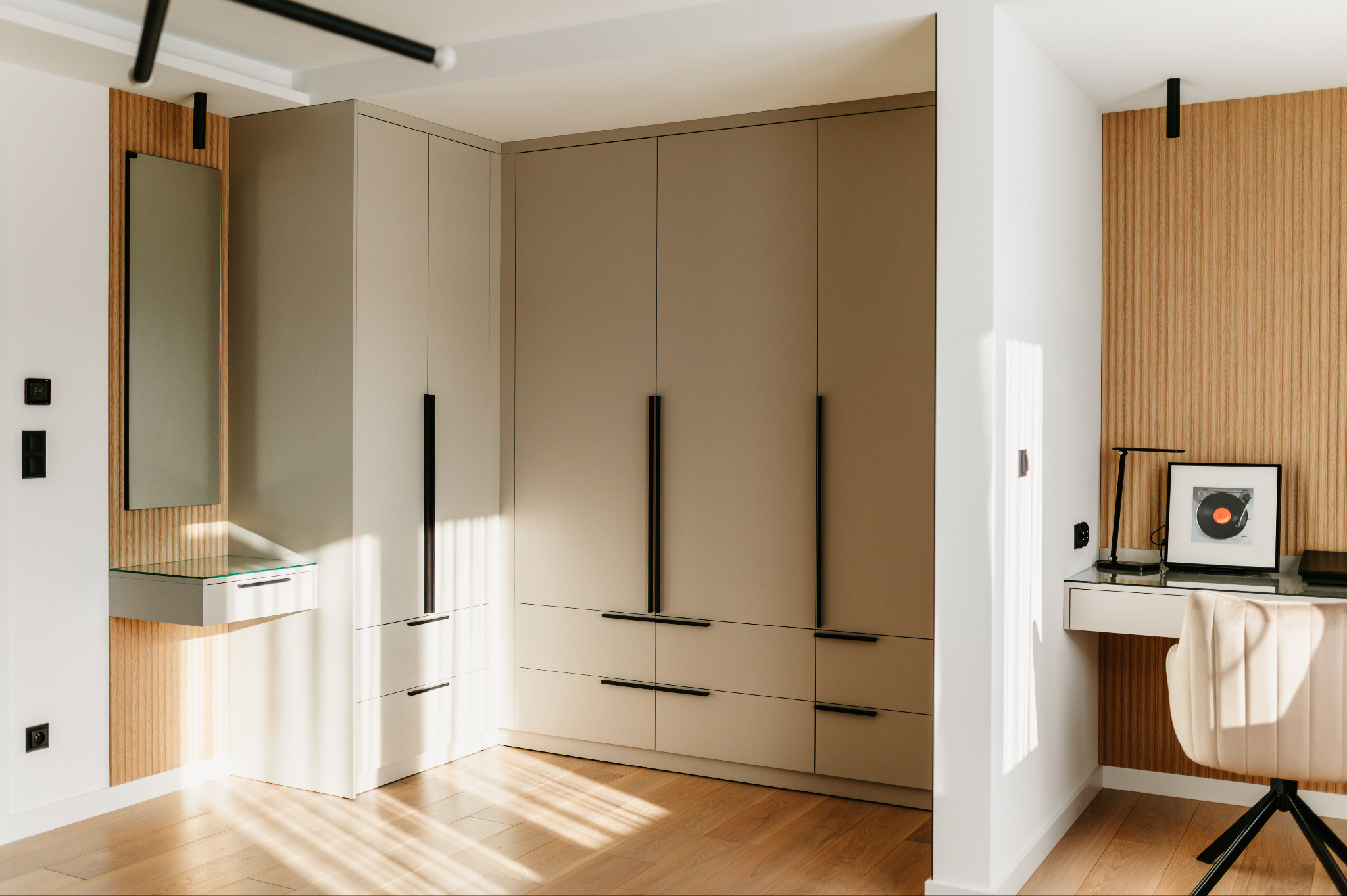 Beżowa zabudowa meblowa z szafą i toaletką z lustrem, uzupełniona biurkiem z lampką i krzesłem na drewnianej podłodze. Ściana wykończona drewnianymi panelami.