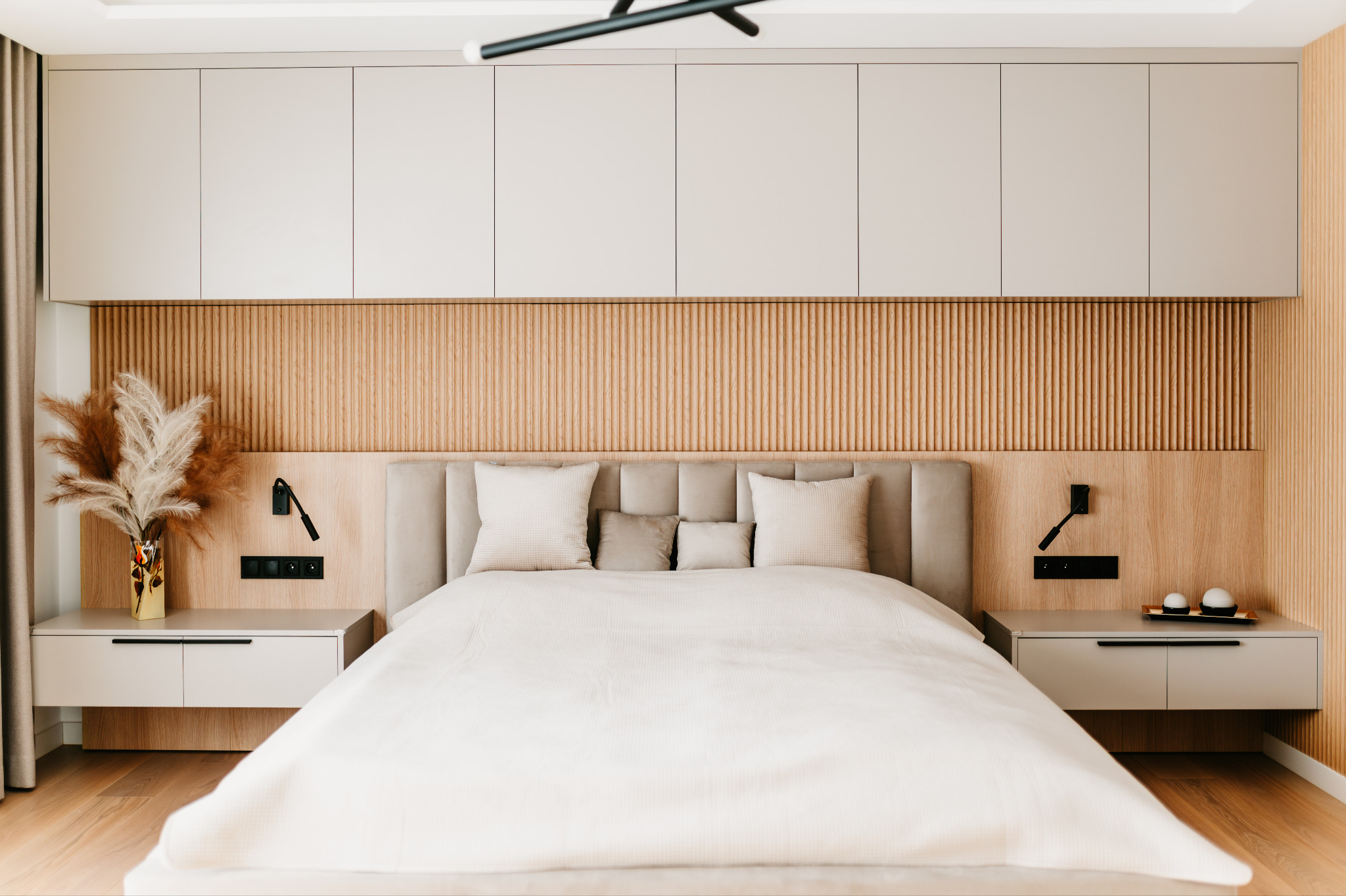 Nowoczesna sypialnia z łóżkiem, drewnianym panelem ściennym i szafkami nocnymi. Minimalistyczny design, stonowana kolorystyka, dekoracyjne trawy w wazonie.