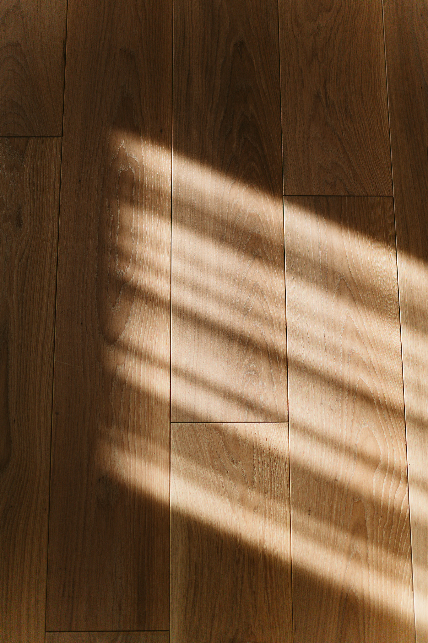 Parkiet z naturalnego drewna w ciepłym odcieniu, oświetlony promieniami słońca tworzącymi geometryczne wzory światła i cienia na powierzchni podłogi.