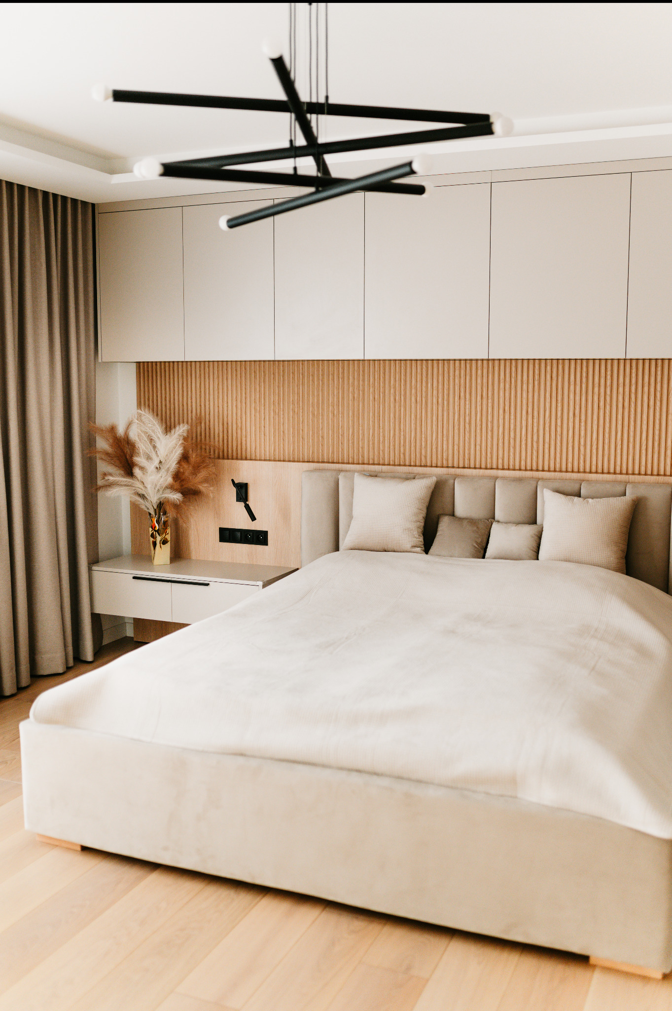 Nowoczesna sypialnia w beżach: łóżko z tapicerowanym zagłówkiem, drewniana lamperia, designerska lampa i minimalistyczne szafki nocne z dekoracją z suszonych traw.