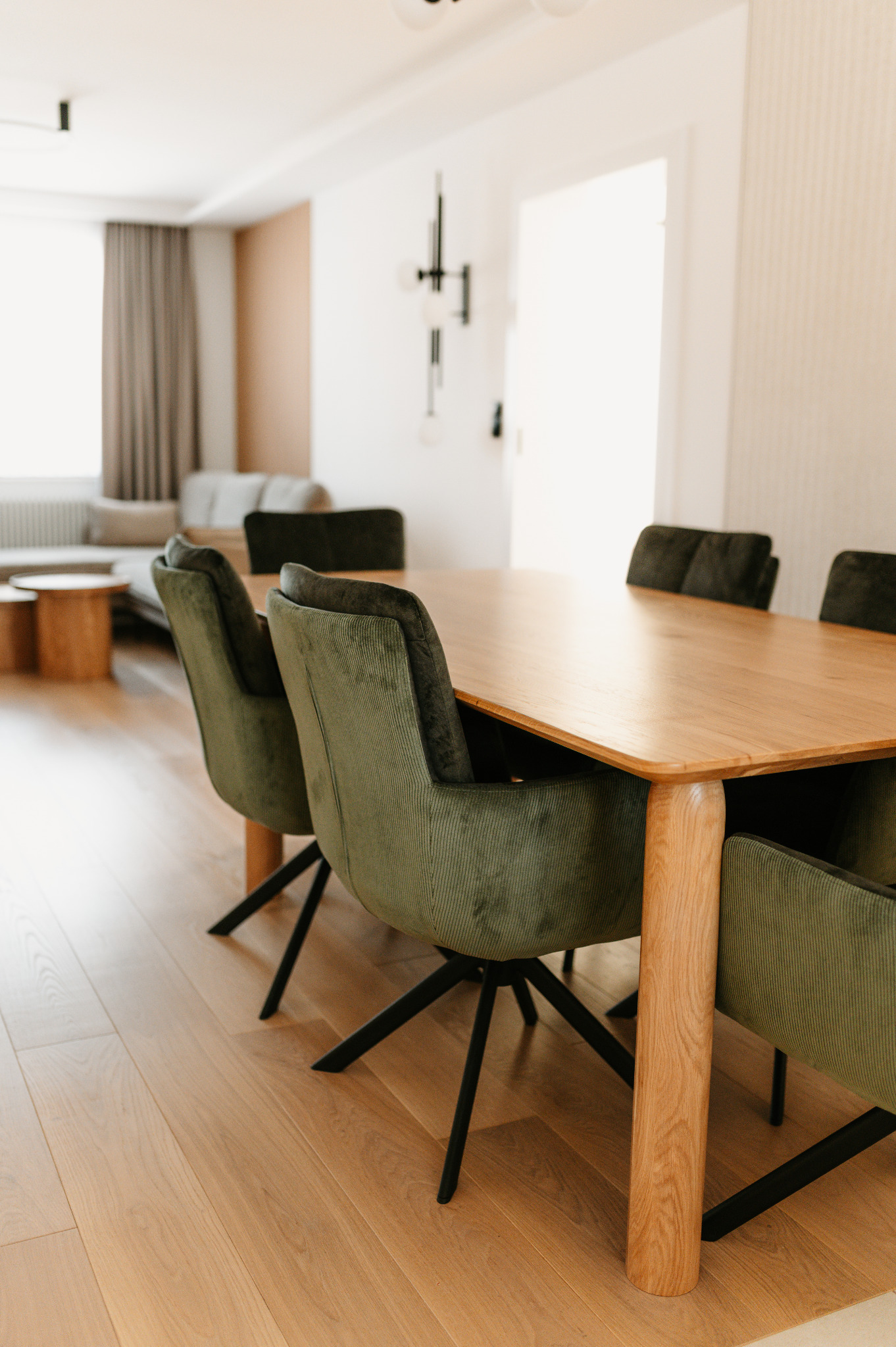 Jasne wnętrze jadalni z drewnianym stołem i zielonymi krzesłami na tle salonu. Minimalistyczny design, parkiet z jasnego drewna, nowoczesne oświetlenie.