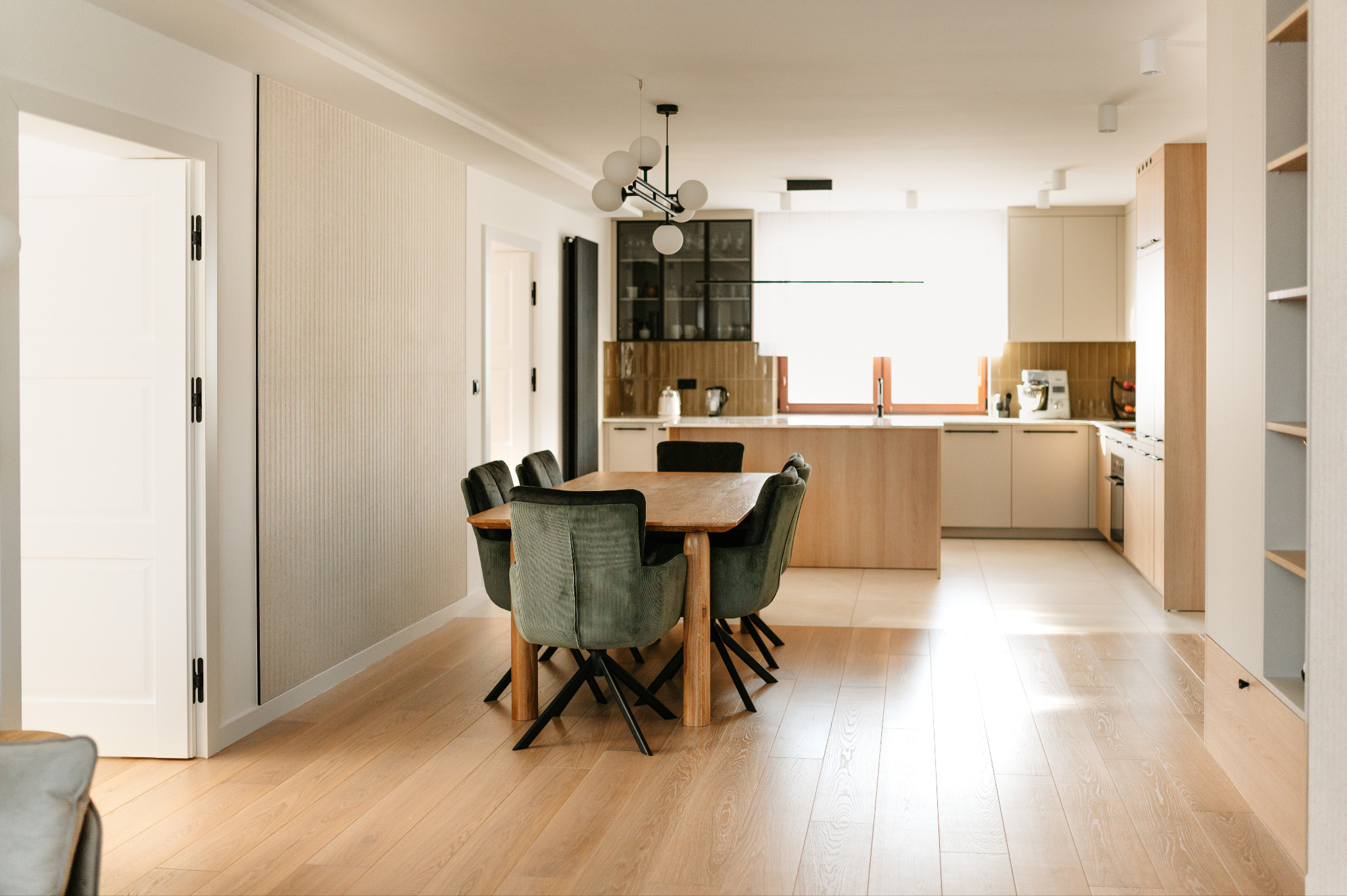 Jasne, przestronne wnętrze z drewnianym stołem i zielonymi krzesłami. W tle otwarta kuchnia z drewnianymi blatami i białymi szafkami. Minimalistyczny design.