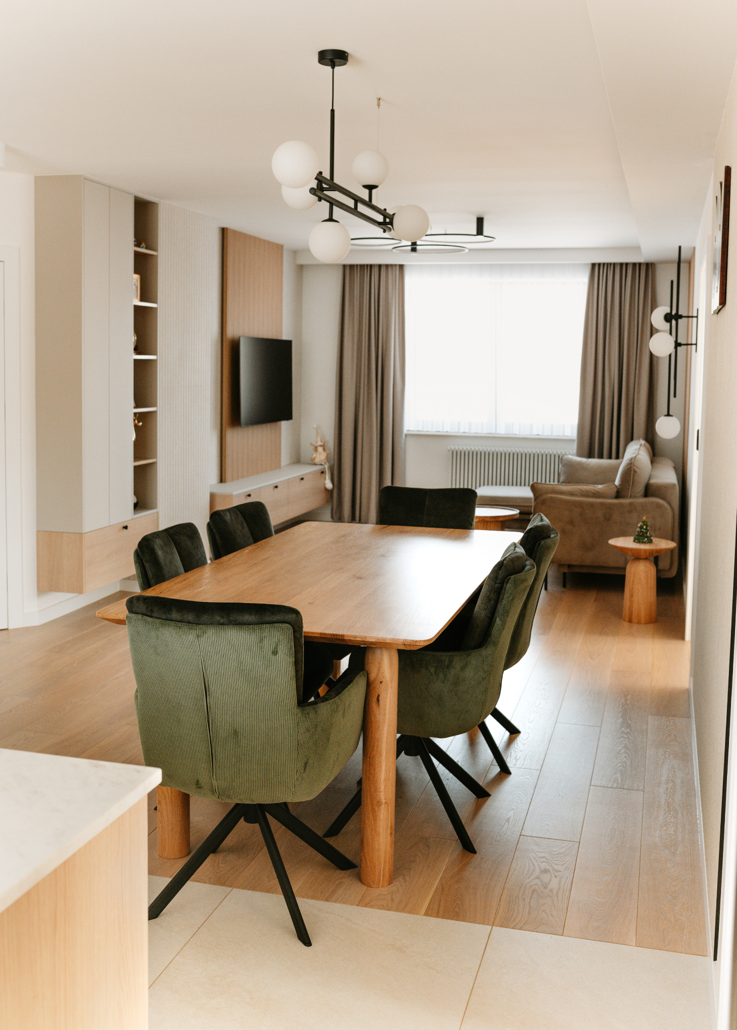 Jasne wnętrze z drewnianym stołem i zielonymi krzesłami. W tle salon z beżową sofą i oknem z zasłonami. Nowoczesne oświetlenie.