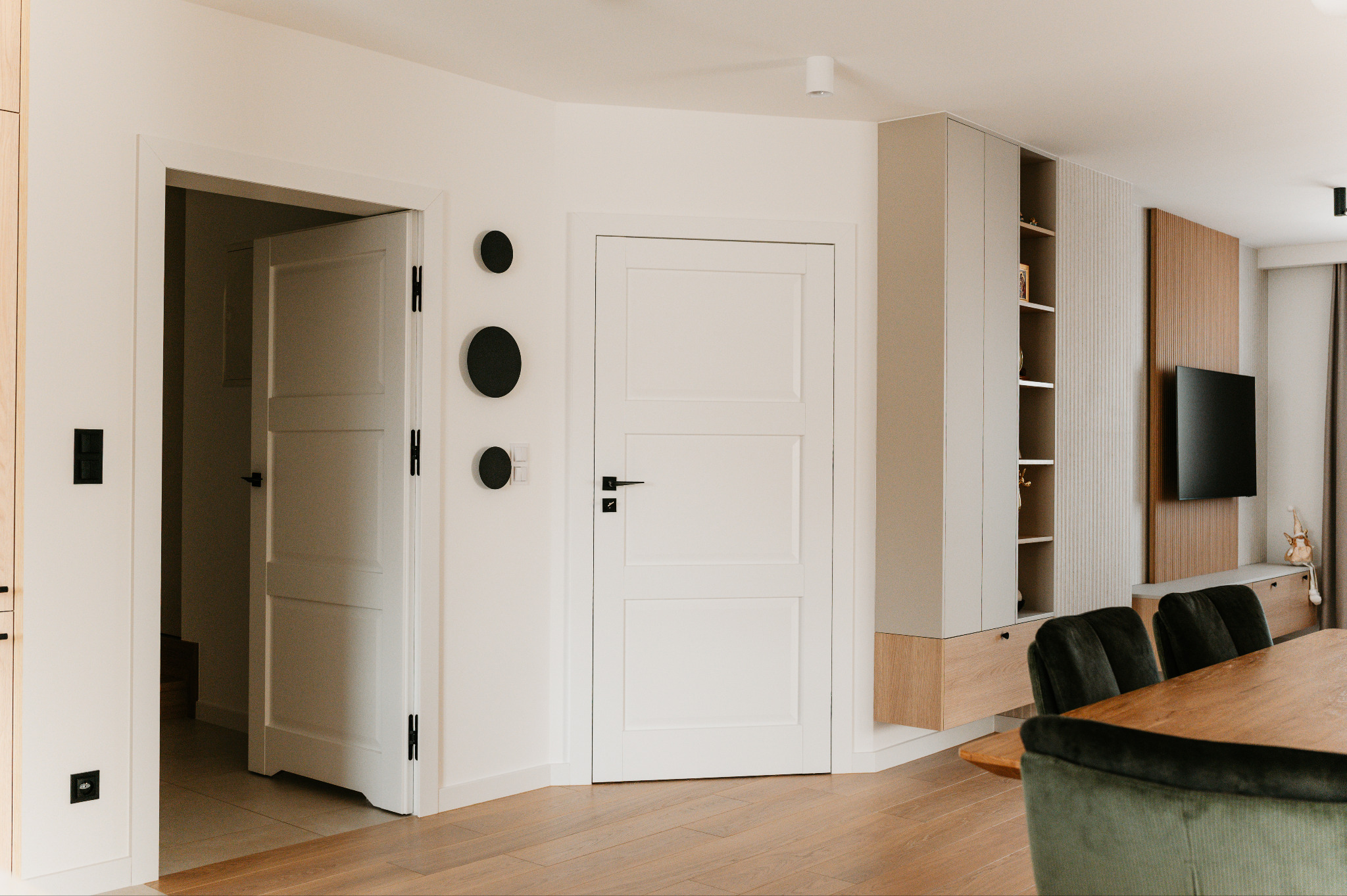 Jasne wnętrze z drewnianą podłogą. Widoczne białe drzwi, zabudowa meblowa z półkami, stół z krzesłami i dekoracyjne, czarne okręgi na ścianie. Minimalistyczny design.