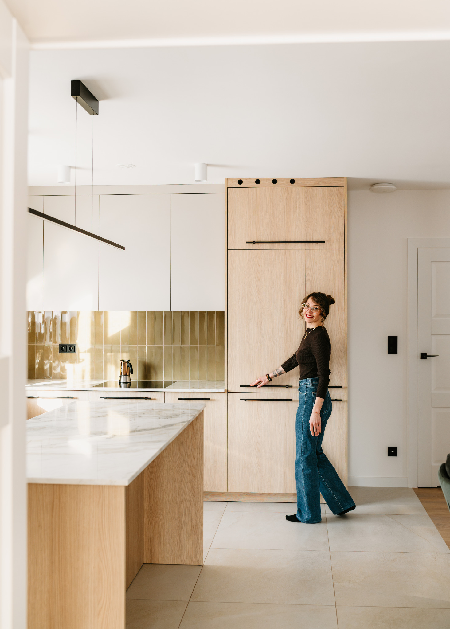 Jasna kuchnia z drewnianymi frontami i marmurowym blatem wyspy. Uśmiechnięta kobieta w okularach stoi na tle zabudowy. Minimalistyczne oświetlenie.