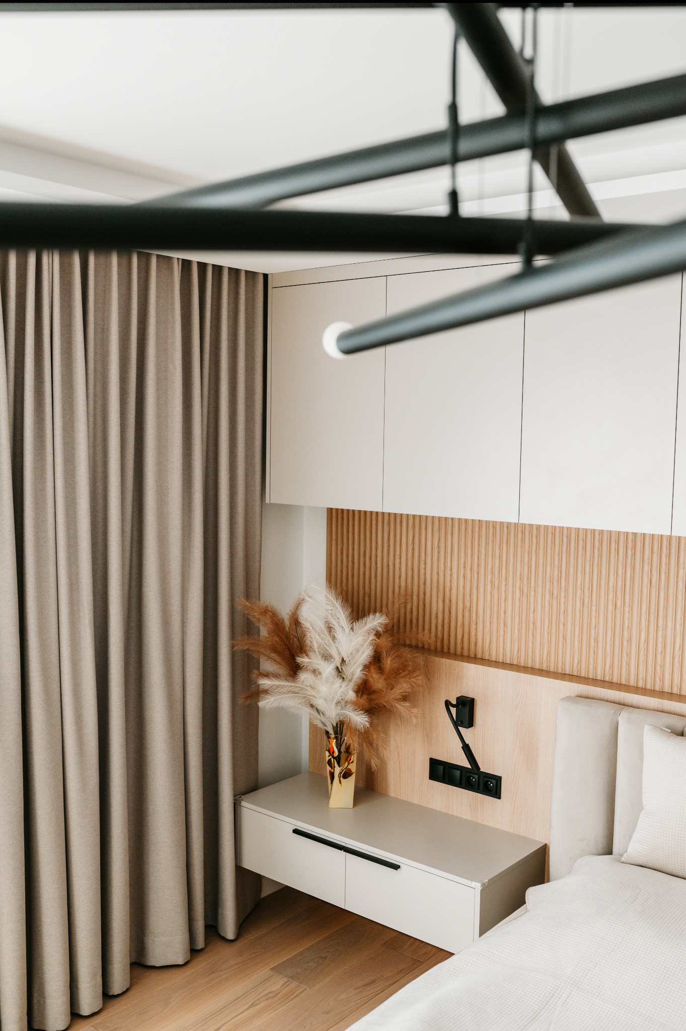 Beżowa sypialnia z drewnianym panelem za łóżkiem, minimalistyczną szafką nocną i dekoracyjnym bukietem w złotym wazonie. Nowoczesne oświetlenie sufitowe.