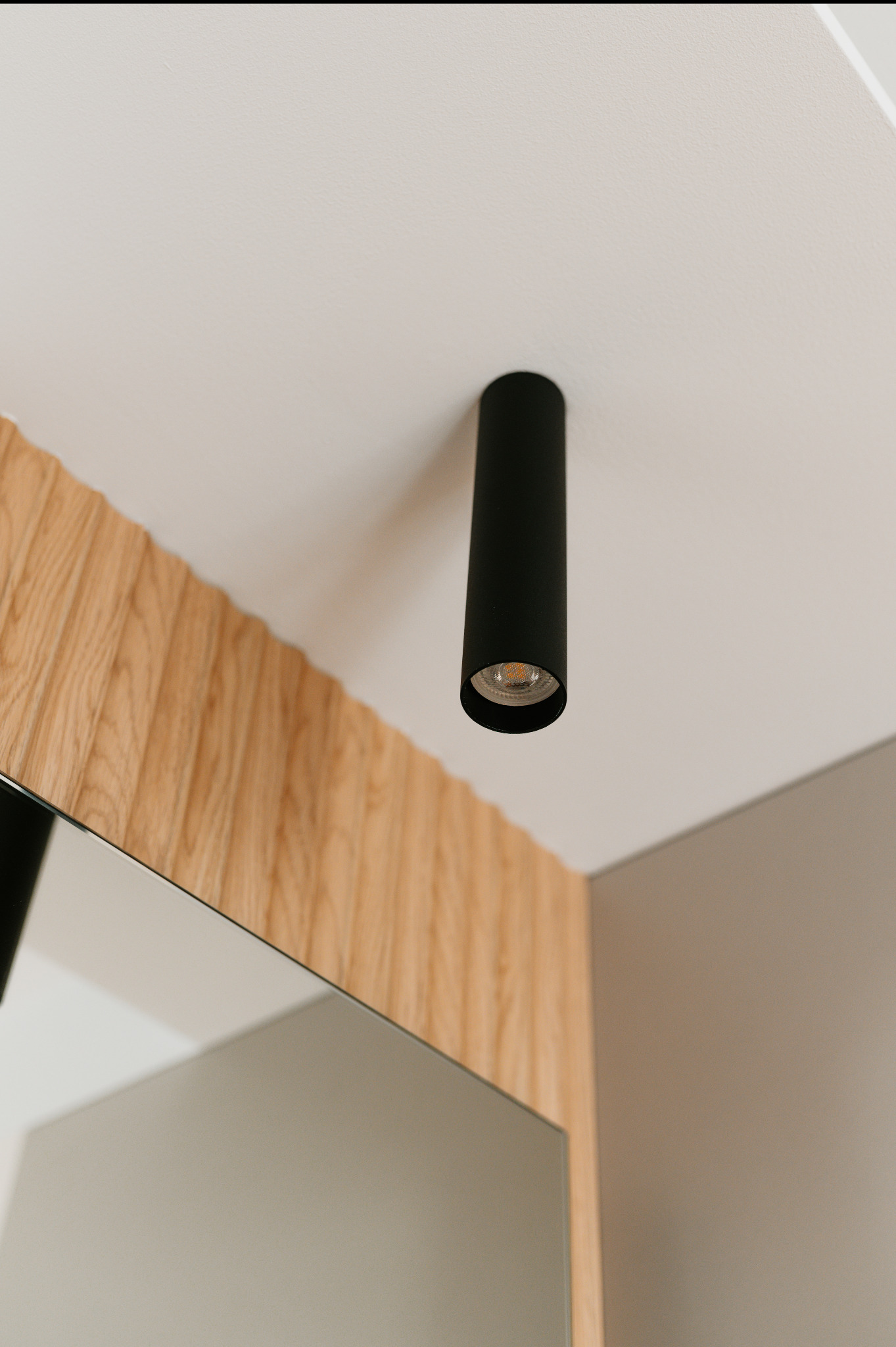 Minimalistyczna łazienka: czarna lampa sufitowa tuba nad lustrem z drewnianą ramą. Nowoczesne oświetlenie punktowe w jasnym wnętrzu.