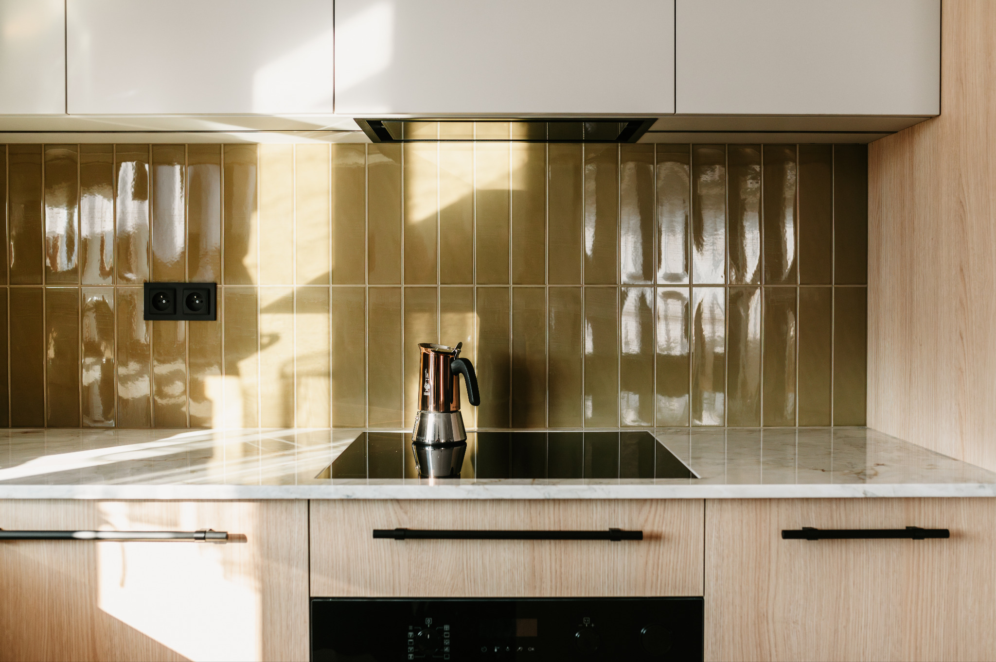Nowoczesna kuchnia z błyszczącymi, oliwkowymi płytkami na ścianie, marmurowym blatem i drewnianymi szafkami. Na płycie indukcyjnej stoi moka.