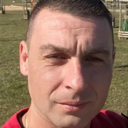 Portret mężczyzny w czerwonej bluzie na tle parku z młodymi drzewkami i błękitnym niebem.