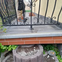 Pracownik w ubrudzonych farbą spodniach stoi na balkonie z dekoracyjną, czarną balustradą, widoczne donice z ziemią i popielniczka z niedopałkami na dole.