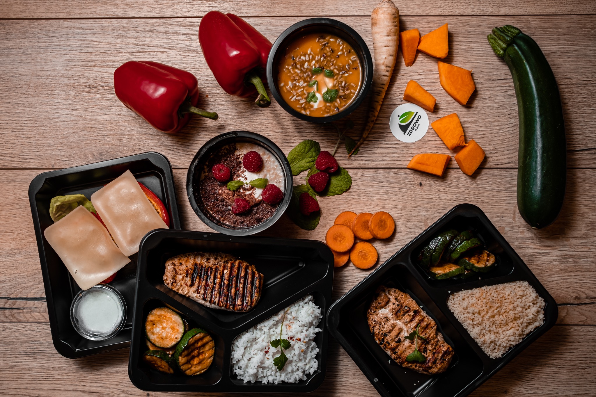 Kompozycja dań dietetycznych w czarnych pojemnikach, zupy, owsianki z malinami, grillowanego mięsa i warzyw, uzupełniona o świeże papryki, marchew, cukinię i logo firmy na drewnianym tle.