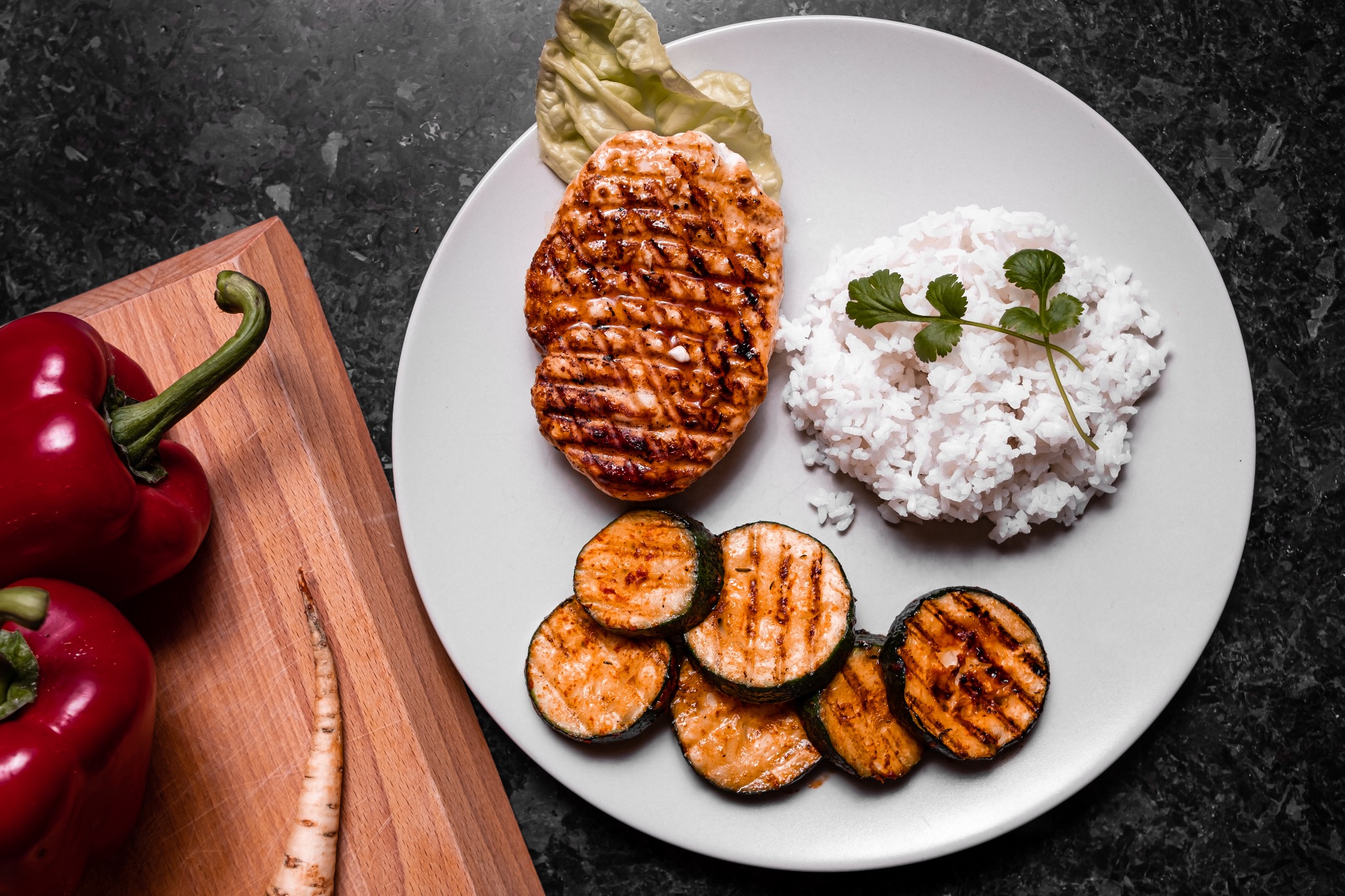 Grillowany filet z kurczaka z ryżem, grillowaną cukinią i sałatą na białym talerzu, z czerwoną papryką i korzeniem pietruszki na drewnianej desce do krojenia, na ciemnym blacie.