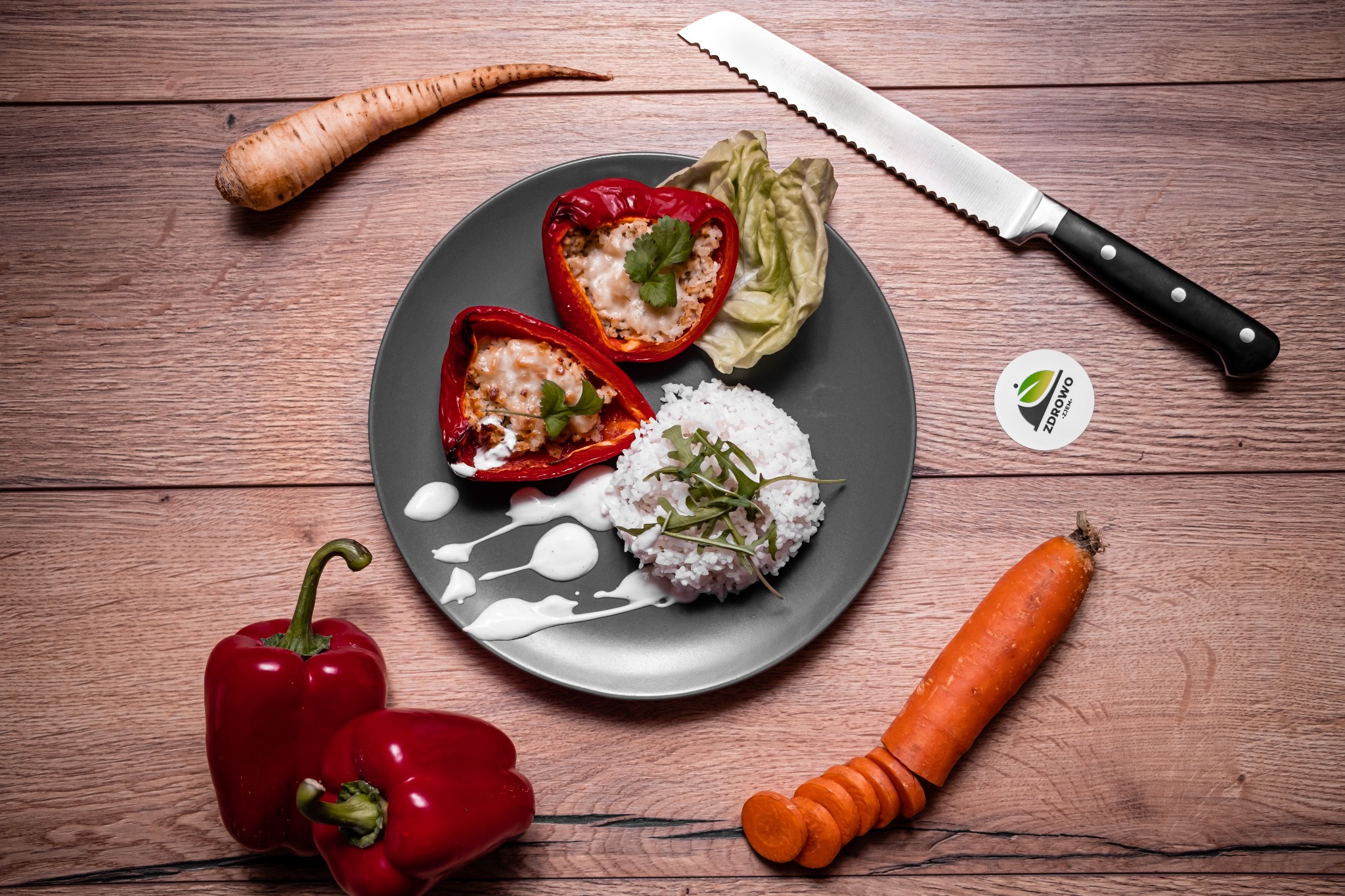 Pełne ujęcie talerza z dwoma połówkami pieczonej czerwonej papryki faszerowanej ryżem i serem, udekorowanego sosem jogurtowym i świeżymi ziołami, obok ryż, sałata, marchewka i pasternak, nóż...
