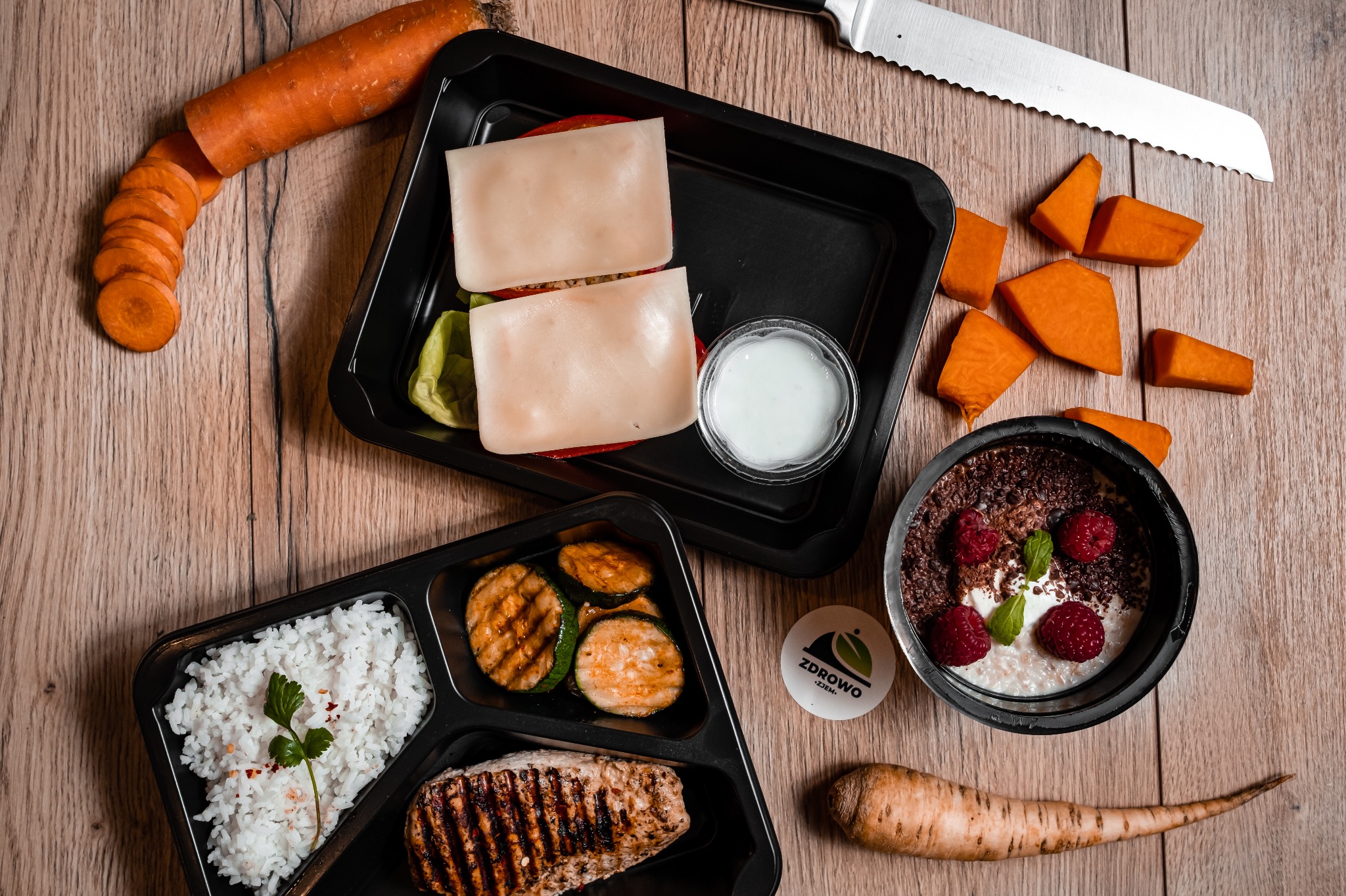 Kompozycja trzech czarnych pojemników z gotowymi daniami: ryż z zielonym listkiem i grillowaną rybą, grillowana cukinia, sałatka z serem, obok miseczka z deserem z malinami, marchew, dynia...