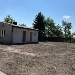 Przygotowany teren pod trawnik, widoczna goła ziemia, budynek gospodarczy z cegły w tle, błękitne niebo.