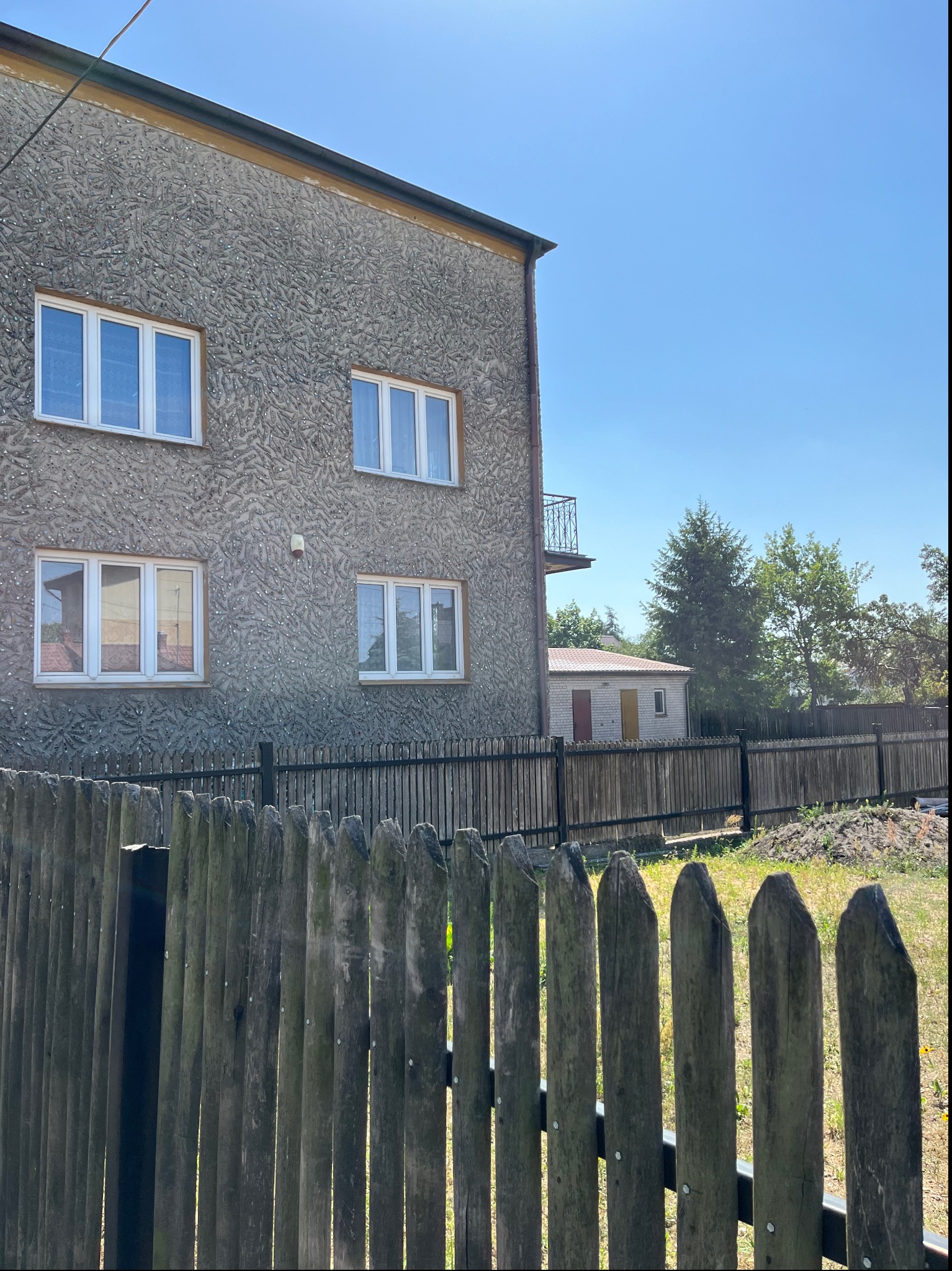 Dwupiętrowy budynek z szarą elewacją widziany zza starego, drewnianego płotu z ostro zakończonymi sztachetami. W tle widoczny mniejszy budynek gospodarczy z czerwonym dachem i fragment drzew.