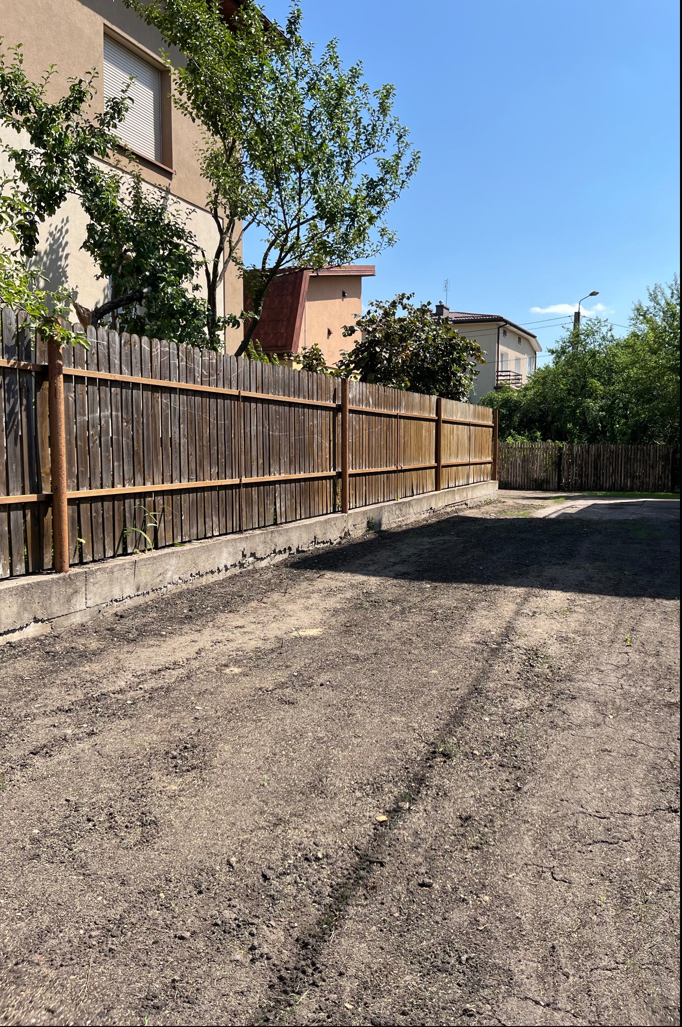 Przygotowany teren pod trawnik, widoczna drewniana brama i ogrodzenie, dom w tle w słoneczny dzień.