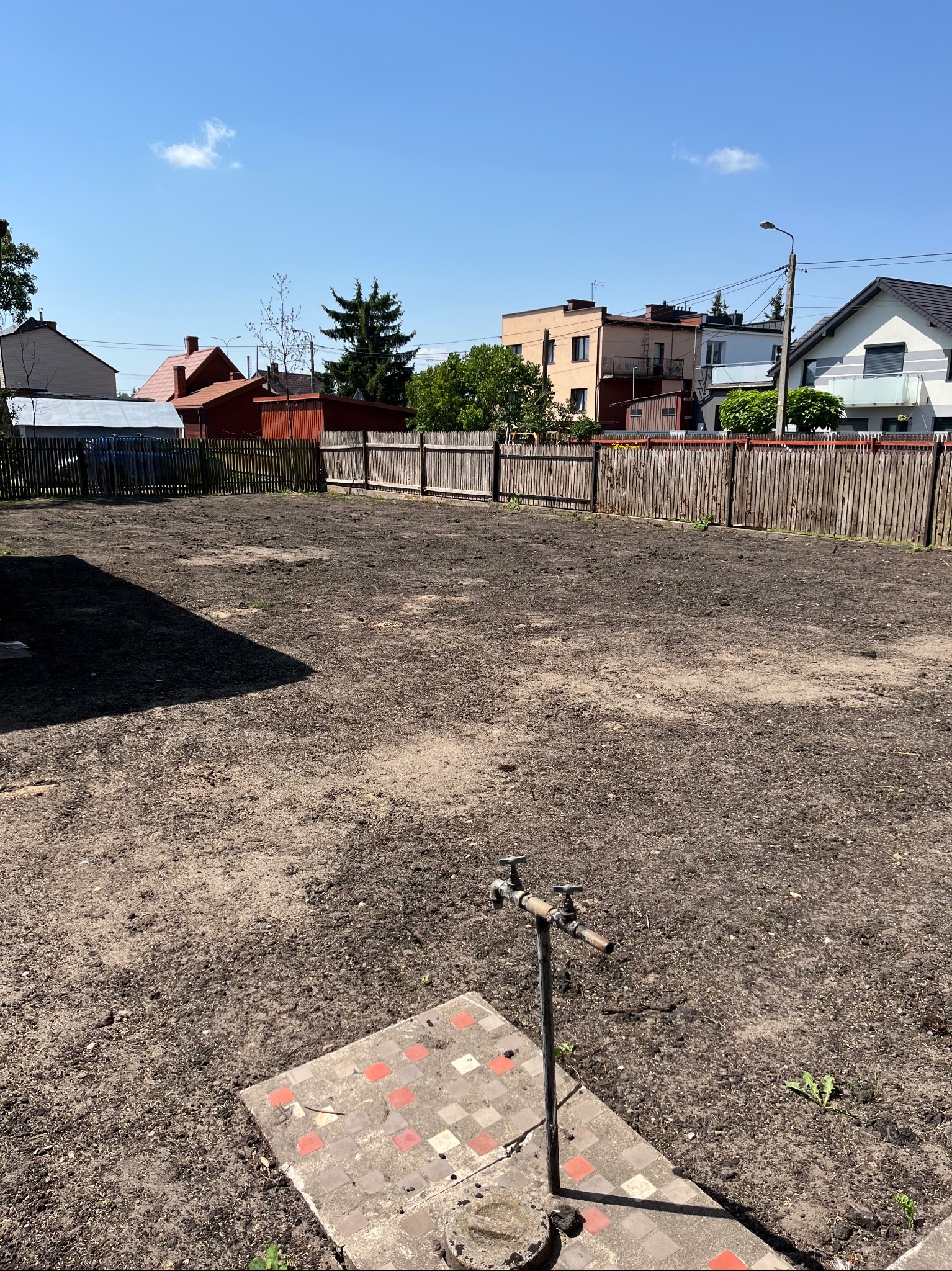 Przygotowany teren pod trawnik z widocznym kranem i płytkami w ogrodzie, ogrodzenie drewniane w tle.