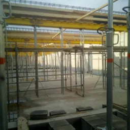 Konstrukcja szalunkowa stropu z żółtymi dźwigarami i metalowymi podporami, widoczne fragmenty muru z bloczków, betonowa posadzka z kałużami.