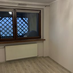 Prywatnie - Jasne wnętrze pokoju po remoncie: panele podłogowe, okno z kratą, grzejnik i ozdobny żyrandol. Ściany pomalowane na jasny odcień.