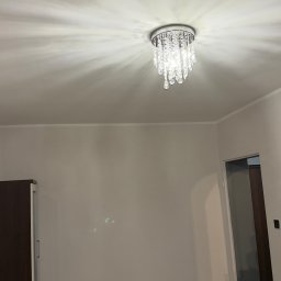 Prywatnie - Jasne wnętrze po odświeżeniu: kremowe ściany, parkiet, ozdobny żyrandol z kryształkami. Widoczny fragment szafy i drzwi. Gniazdko elektryczne na ścianie.