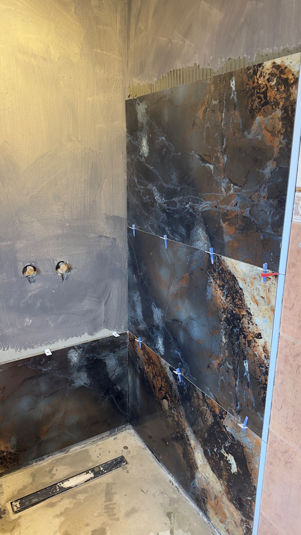 Fragment łazienki w trakcie wykańczania: ciemne, marmurkowe płytki na ścianie z niebieskimi krzyżykami dystansowymi, surowa ściana z otworami na instalacje.